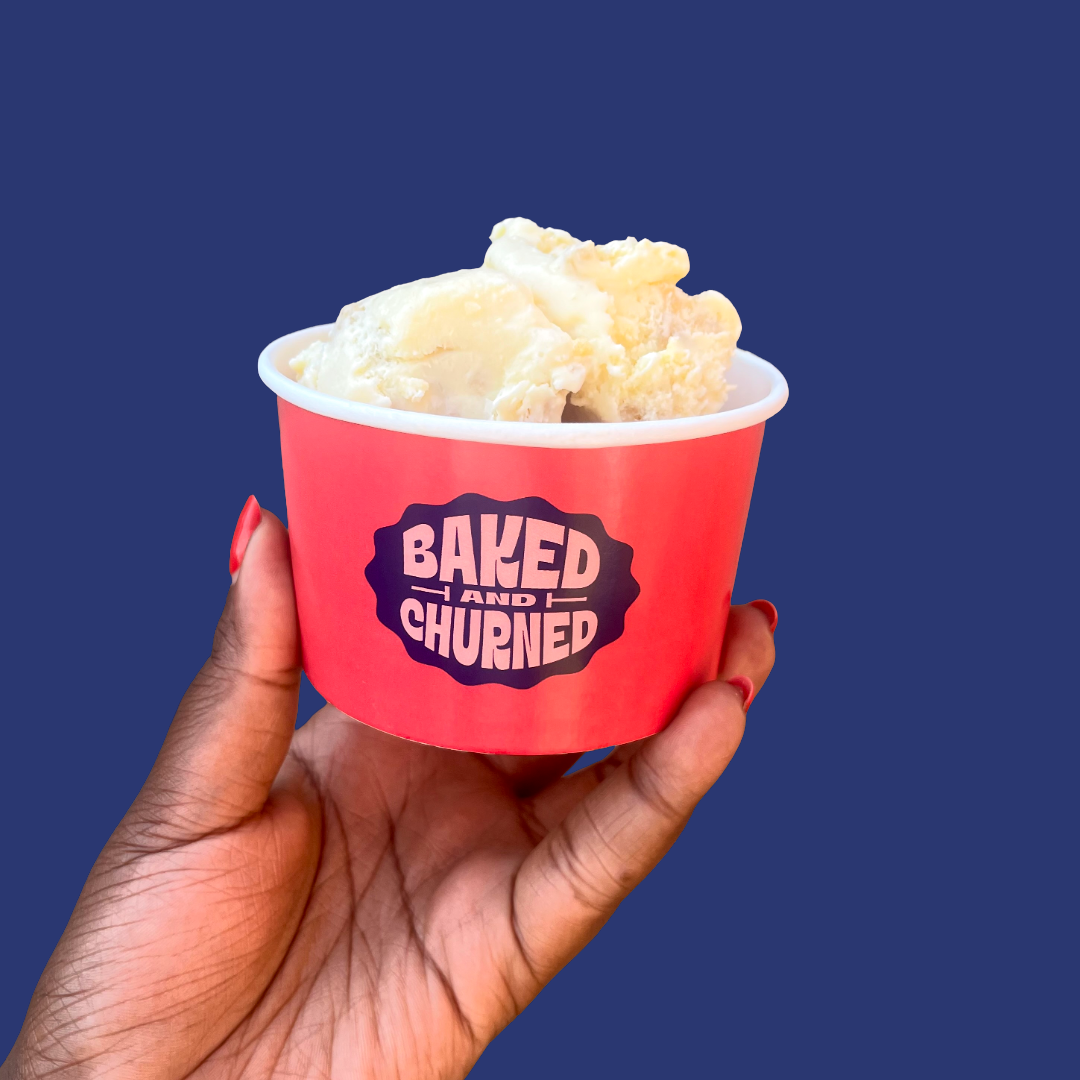 A hand holding a cup of vanilla ice cream in a pink container with a blue background. The cup has the logo 'Baked and Churned'.