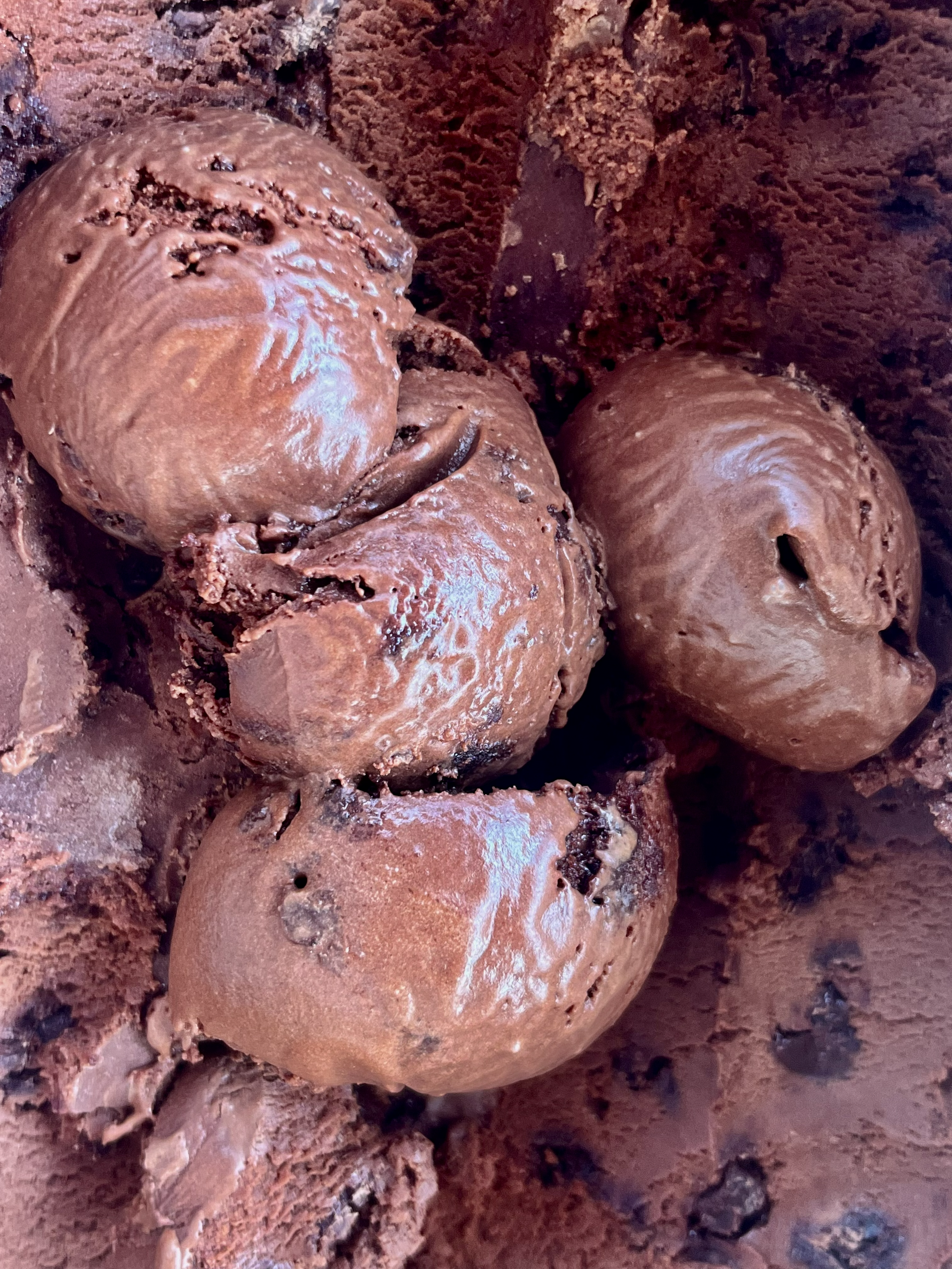 Close-up of chocolate ice cream scoops with a creamy, smooth texture, resting on a bed of chocolate brownie bark chunks.