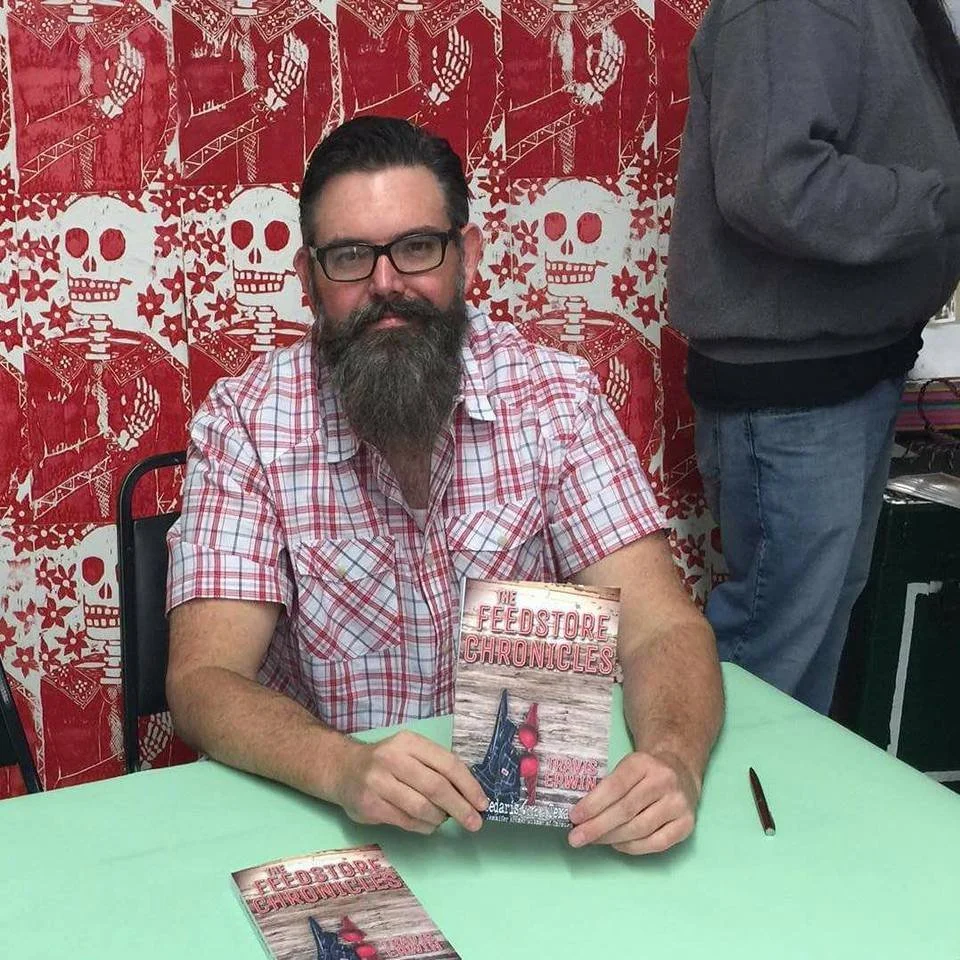 Travis Erwin sitting at a table, holding a book titled 'The Feedstore Chronicles'. The background features a red and white wallpaper pattern of skulls and skeletons.