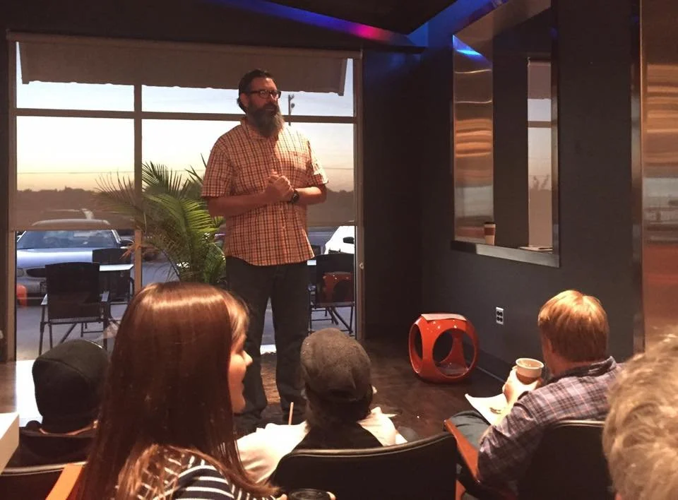 Travis Erwin stands and speaks to a group of people seated indoors during sunset. Large windows show parked cars and a sunset outside.