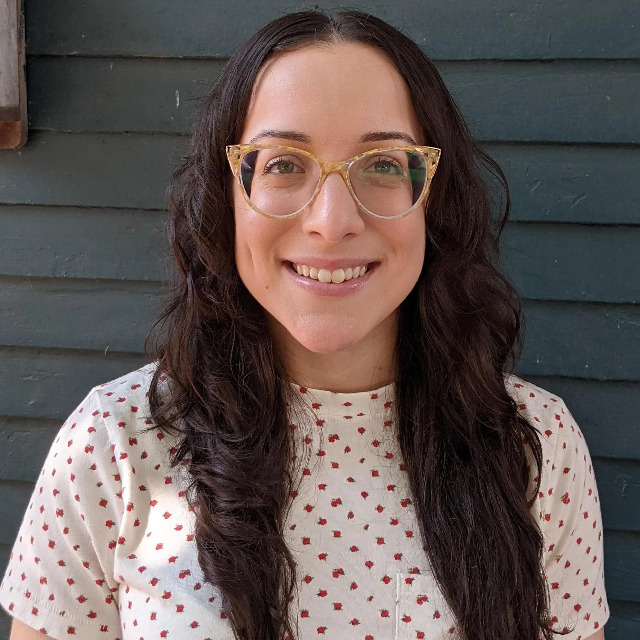 A woman with long dark curly hair wearing glasses and a white shirt with small red polka dots standing in front of a dark wooden wall.