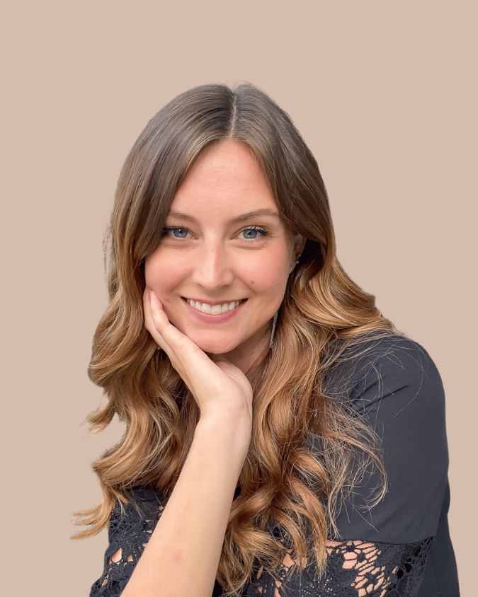 A woman with long, wavy, light brown hair smiling with her head resting on her hand against a beige background.