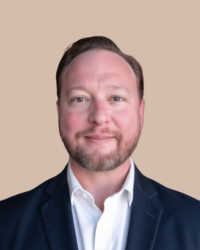 Headshot of a man with light skin, light brown hair, a beard, wearing a dark suit jacket and a white shirt, smiling, with a plain beige background.