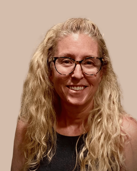A woman with long, curly blonde hair, wearing glasses and a black top, smiling at the camera.