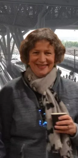 A woman smiling outdoors near a body of water with a bridge structure overhead.