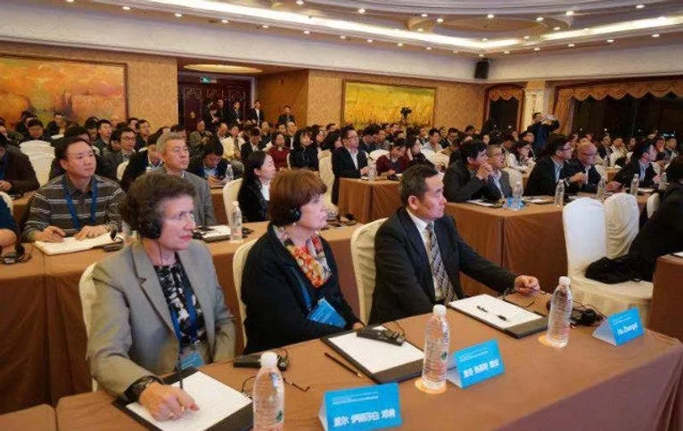 Large conference room filled with people seated at tables, listening to a speaker. The front row has three individuals with name tags and bottled water. The room has a decorated ceiling and paintings on the walls.