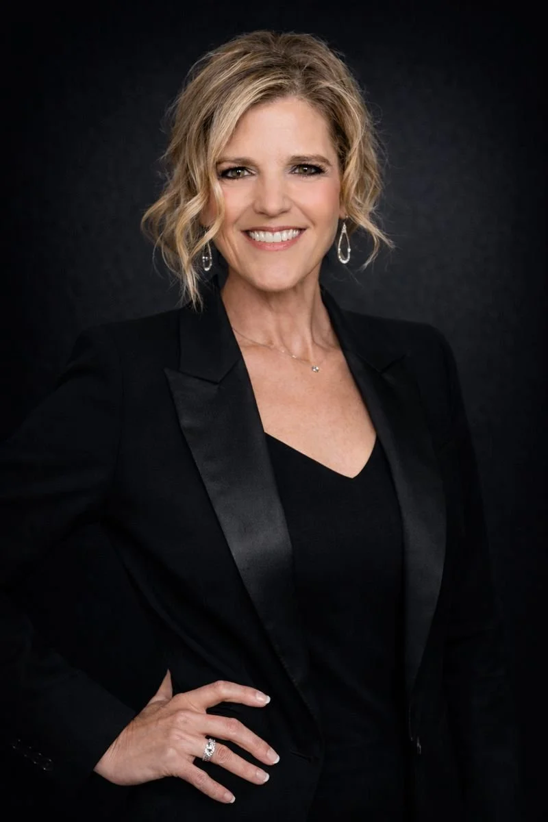 A woman with short curly blonde hair, wearing a black blazer with satin lapels, black top, and silver jewelry, smiling against a dark background.