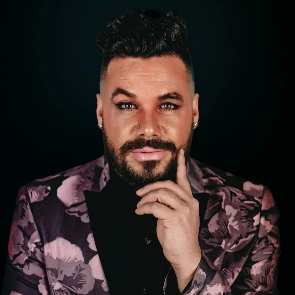 Portrait of a man with short, styled dark hair, a beard, and piercing eyes, wearing earrings and a patterned blazer, against a dark background.