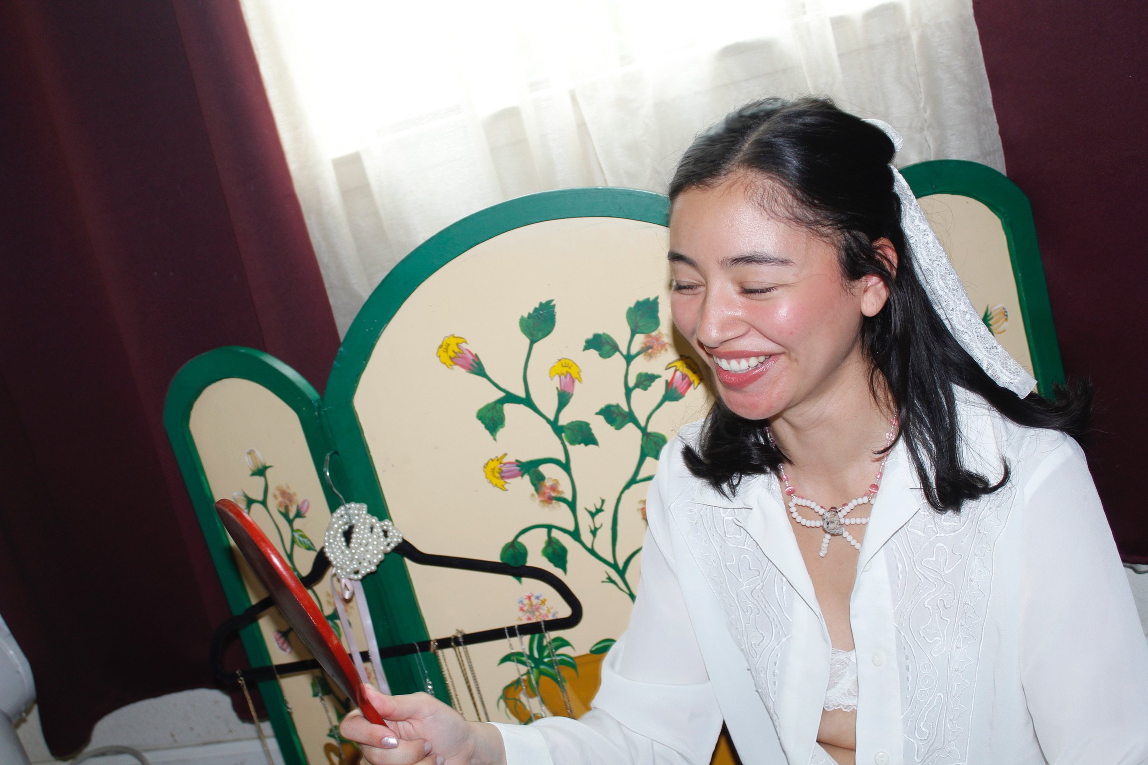 A woman in a white blouse smiling as she looks at her phone, sitting in front of a decorative folding screen with floral artwork.