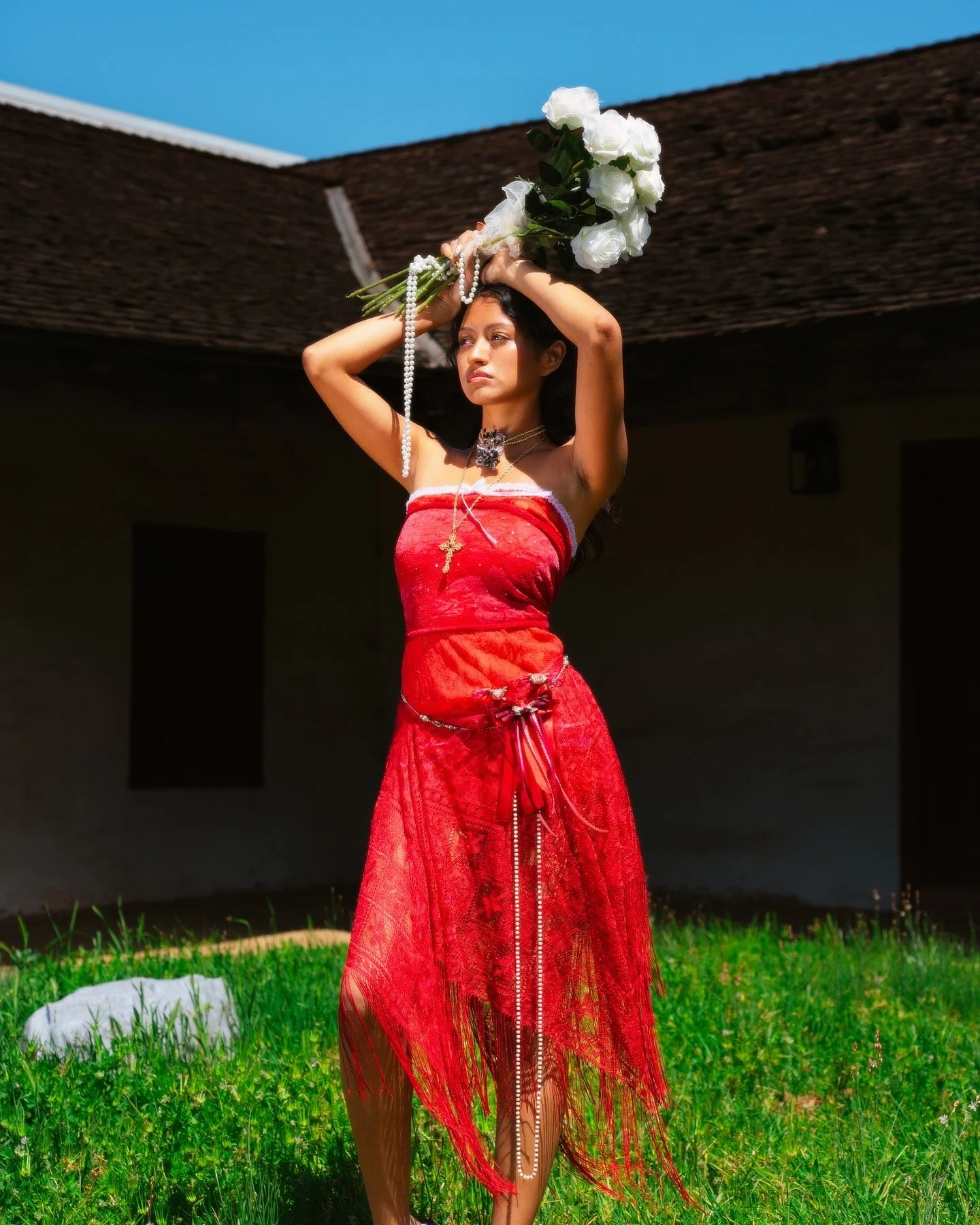 A woman in a red dress stands outdoors on grass, holding a bouquet of white roses above her head, with a building and clear blue sky in the background.