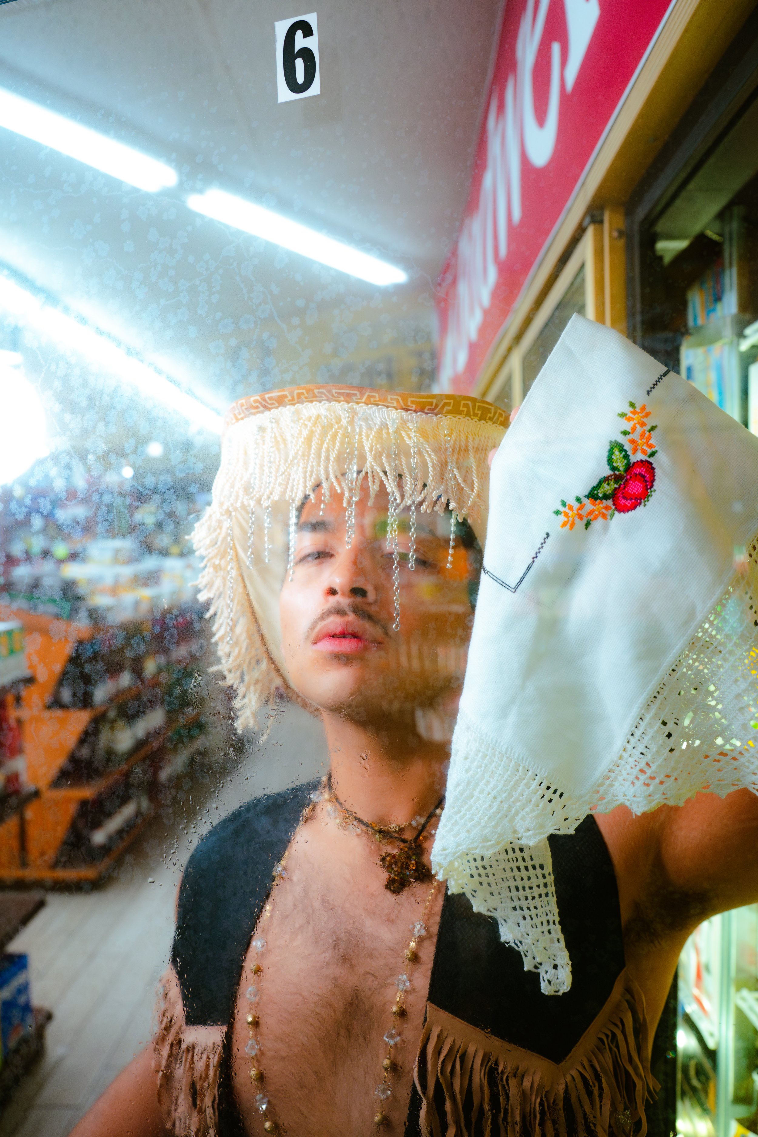 A man wearing a straw sombrero costumed with beads and dangling ornaments, looking through a rainy store window while cleaning it with a white cloth embroidered with berries and flowers. The store appears to be a bookstore or convenience store.
