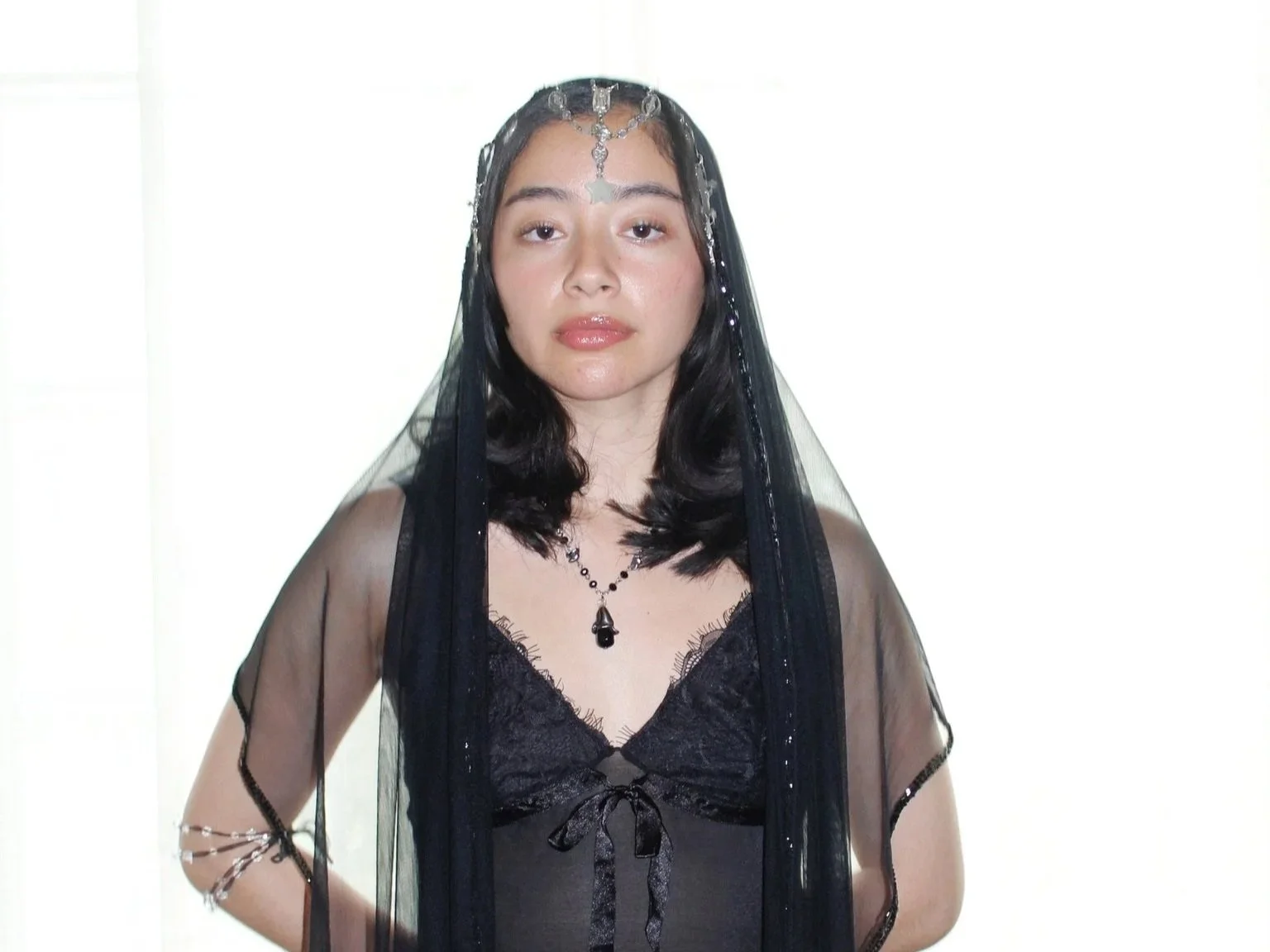 A woman with dark hair in a black lace top, layered necklaces, and a sheer black veil with silver accents, standing against a white background.