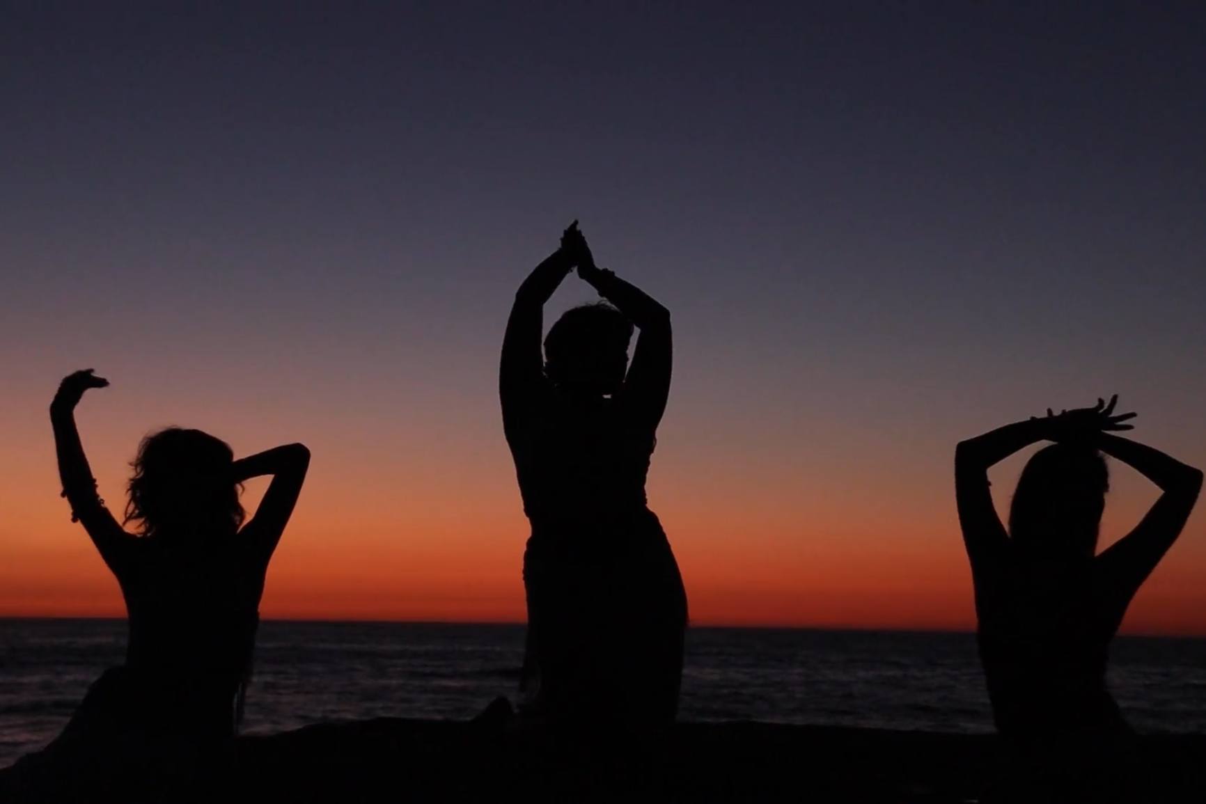 Silhouettes of three people practicing yoga outdoors during sunset by the ocean.