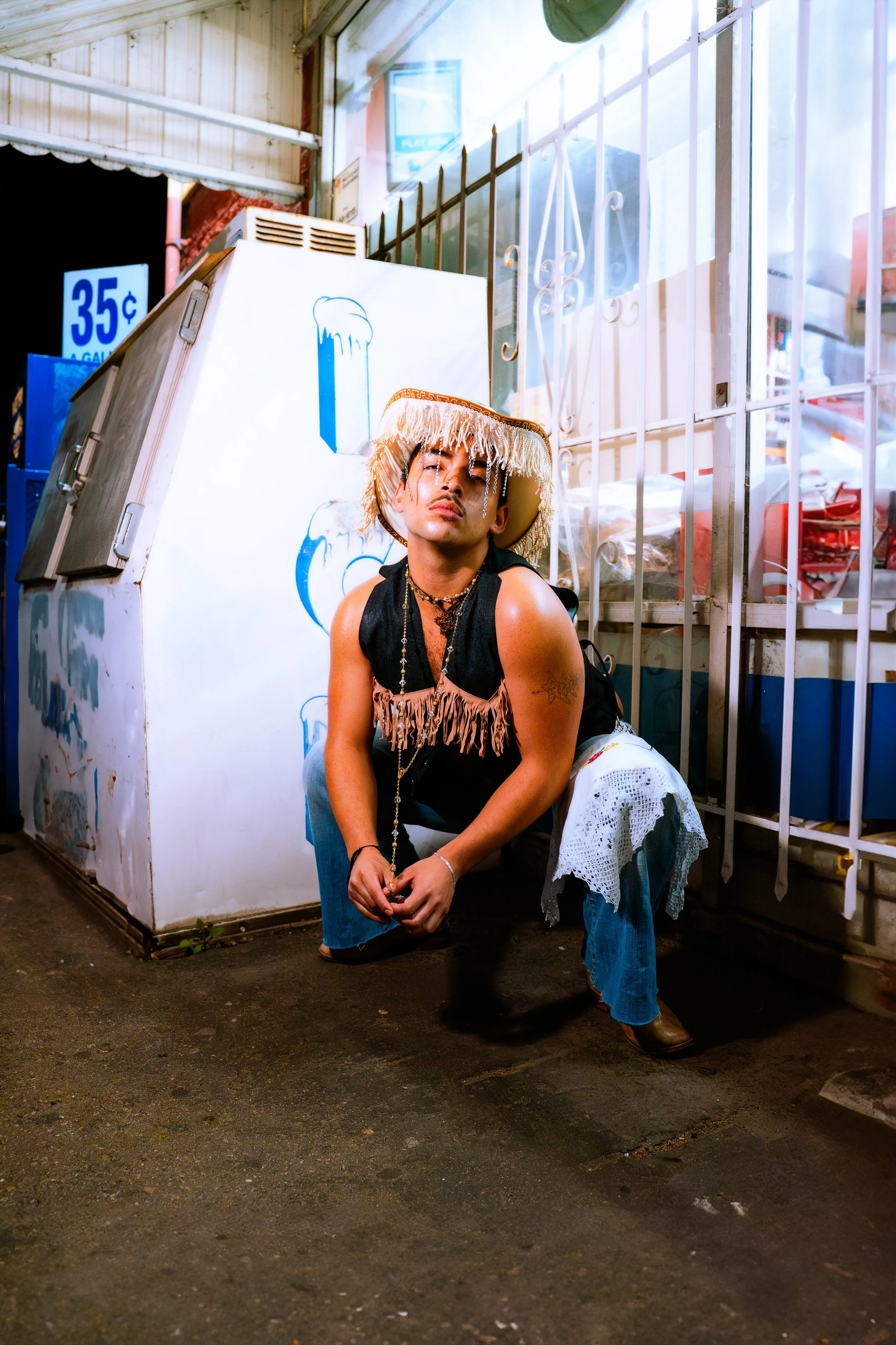 A person wearing a fringed vest, blue jeans, cowboy hat, and jewelry squatting on the ground outside a storefront. They have a tattoo on their arm and are positioned next to an ice machine with water stains and a sign in the background.