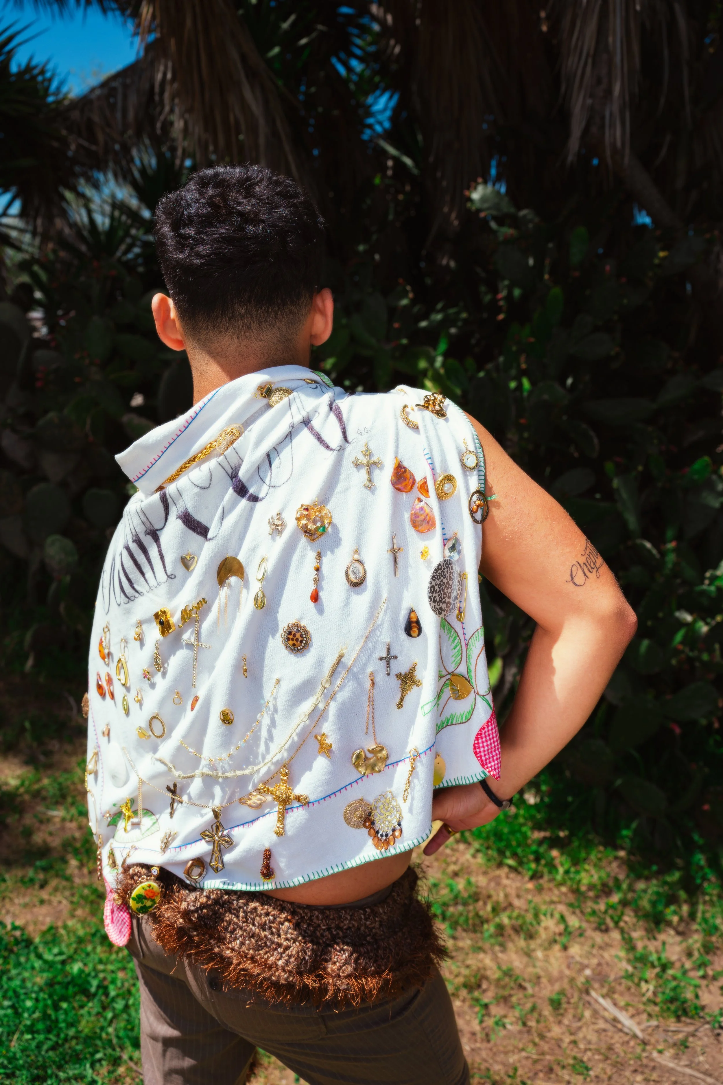 Person with short hair, wearing a white garment decorated with various colorful embroidered jewelry and religious symbols, standing outdoors near green foliage under a blue sky.