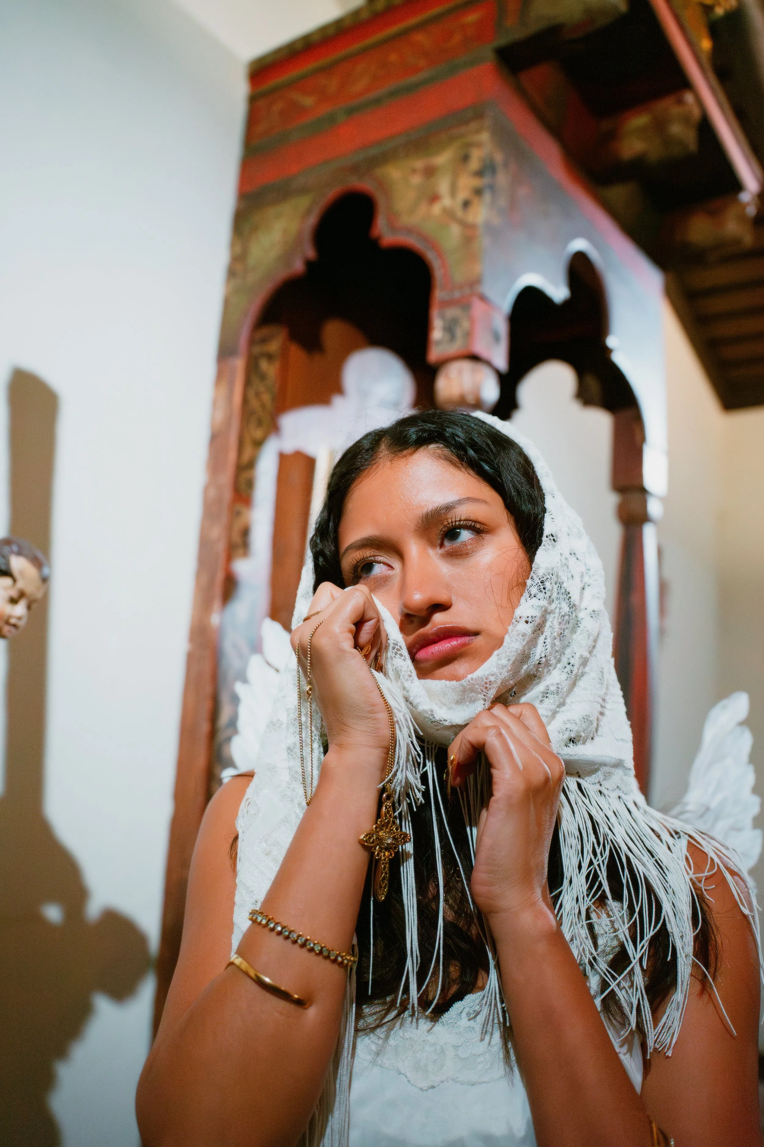 A woman wearing a lace headscarf and gold jewelry, looking thoughtfully, standing in a room with an ornate wooden structure behind her.