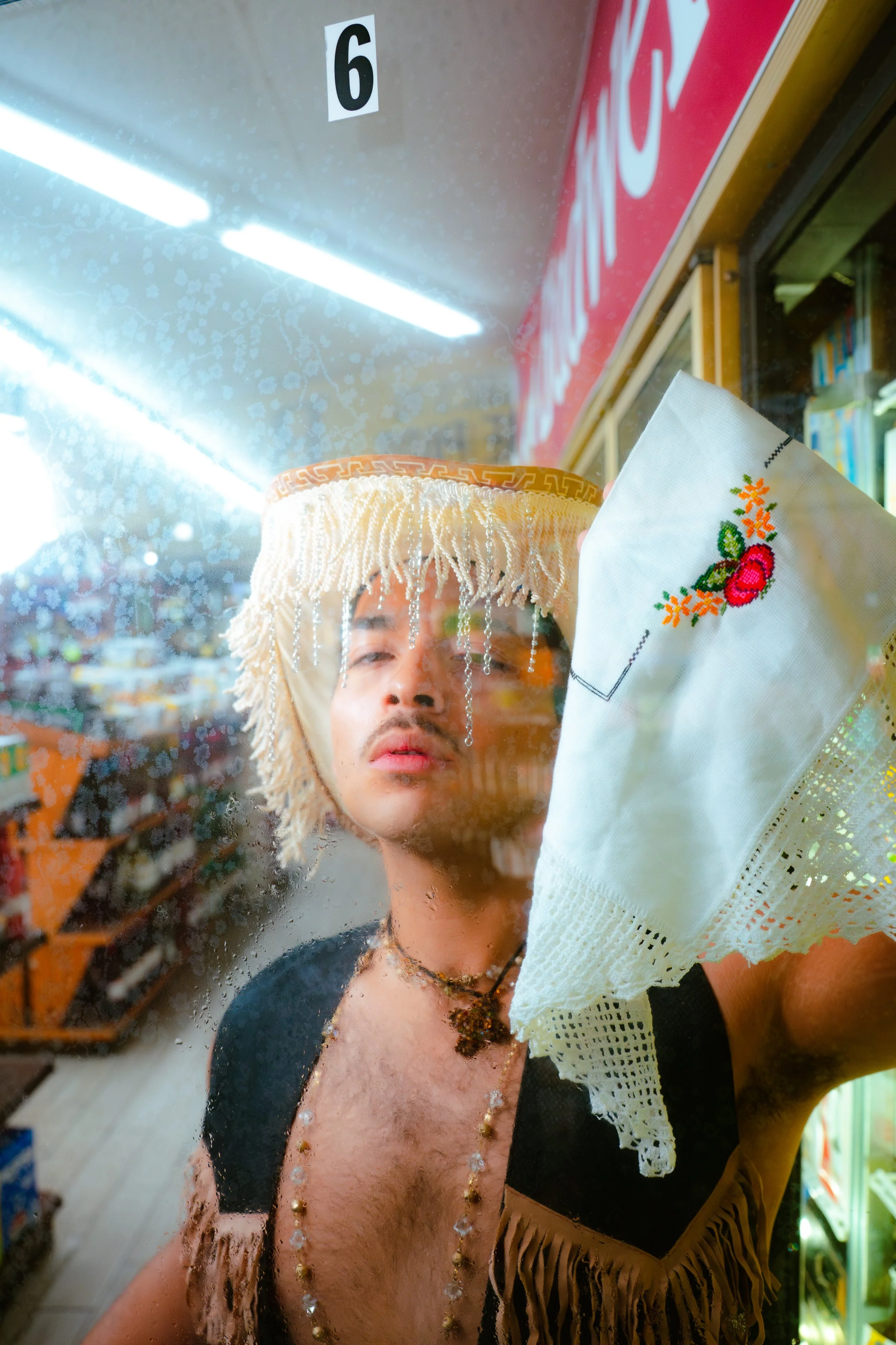 A young man looking through a glass window with water droplets, wearing a hat with fringe, necklaces, and a dark shirt.
