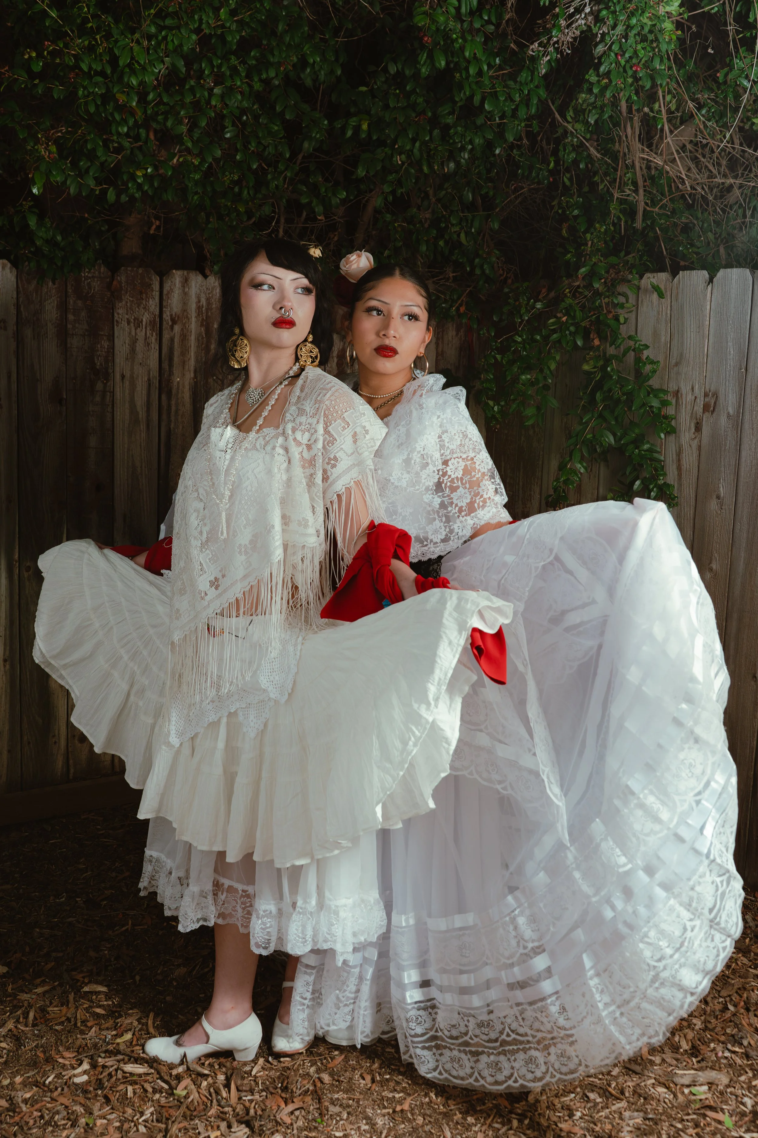 Two women in vintage white lace dresses with red accents, sitting outdoors in front of a wooden fence and greenery, posed for a photoshoot.