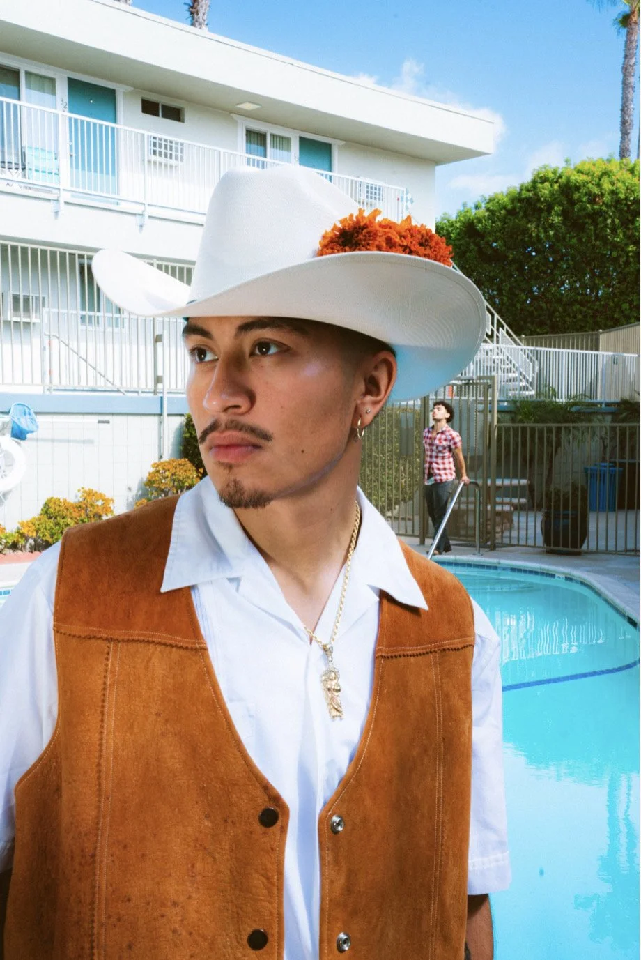 Young man wearing a white cowboy hat decorated with orange flowers, white shirt, brown vest, and gold chain with pendant, standing by a swimming pool with apartment complex and trees in the background.