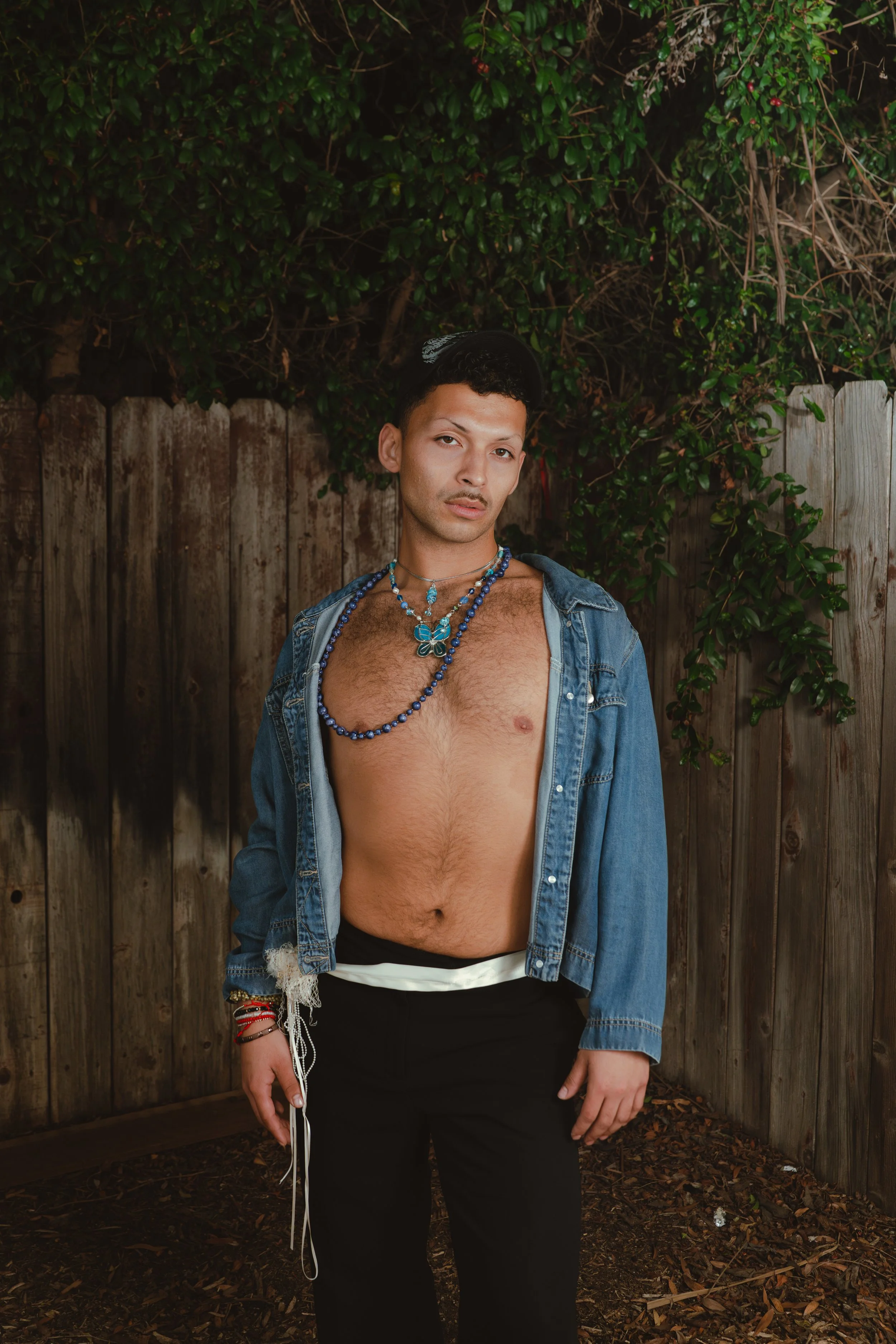 A young man standing outdoors in front of a wooden fence and green foliage, wearing a denim jacket, multiple necklaces, and black pants, with a serious expression.