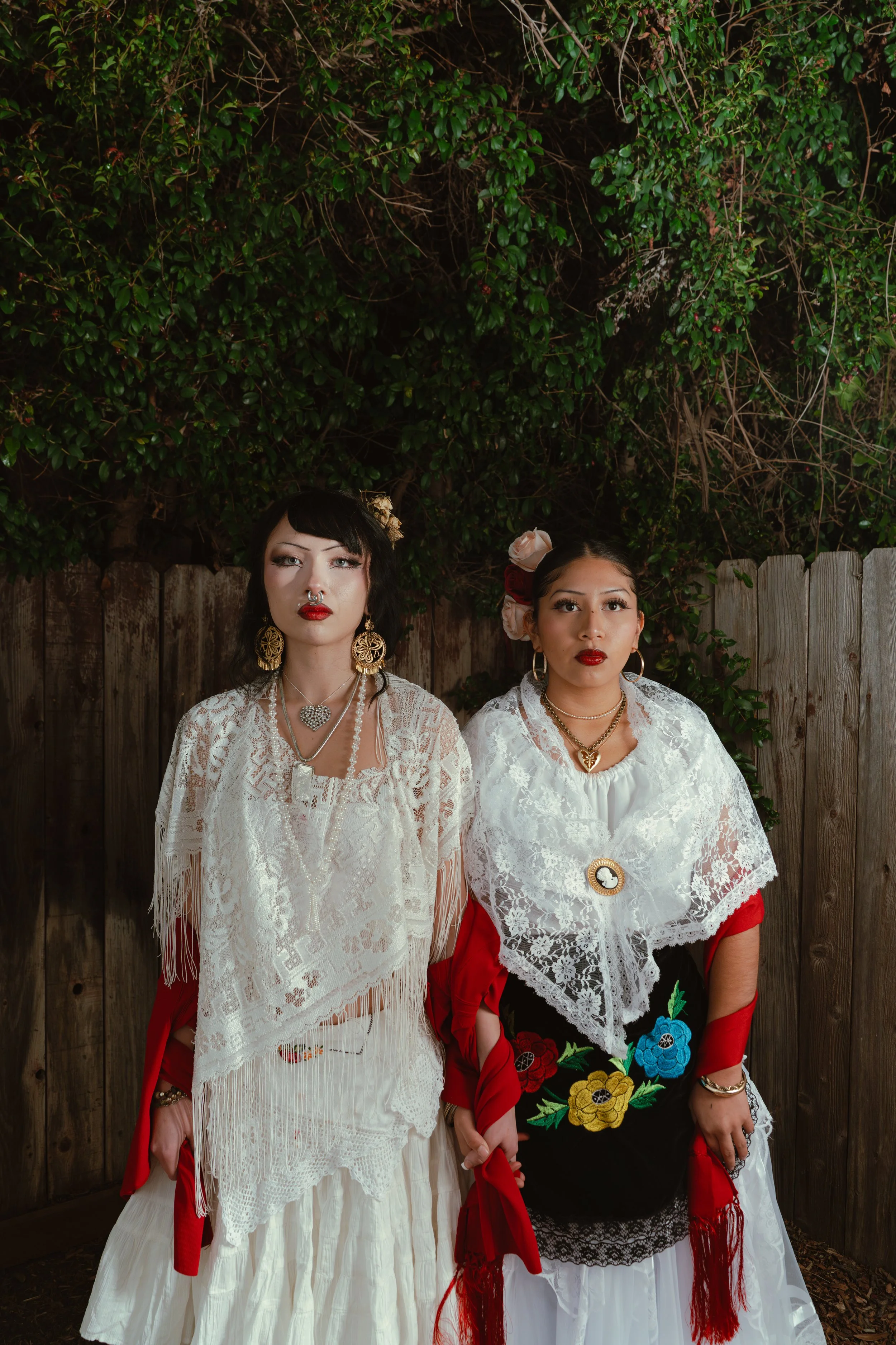 Two women dressed in traditional Mexican attire with white lace shawls and embroidered skirts, standing in front of a wooden fence and greenery.