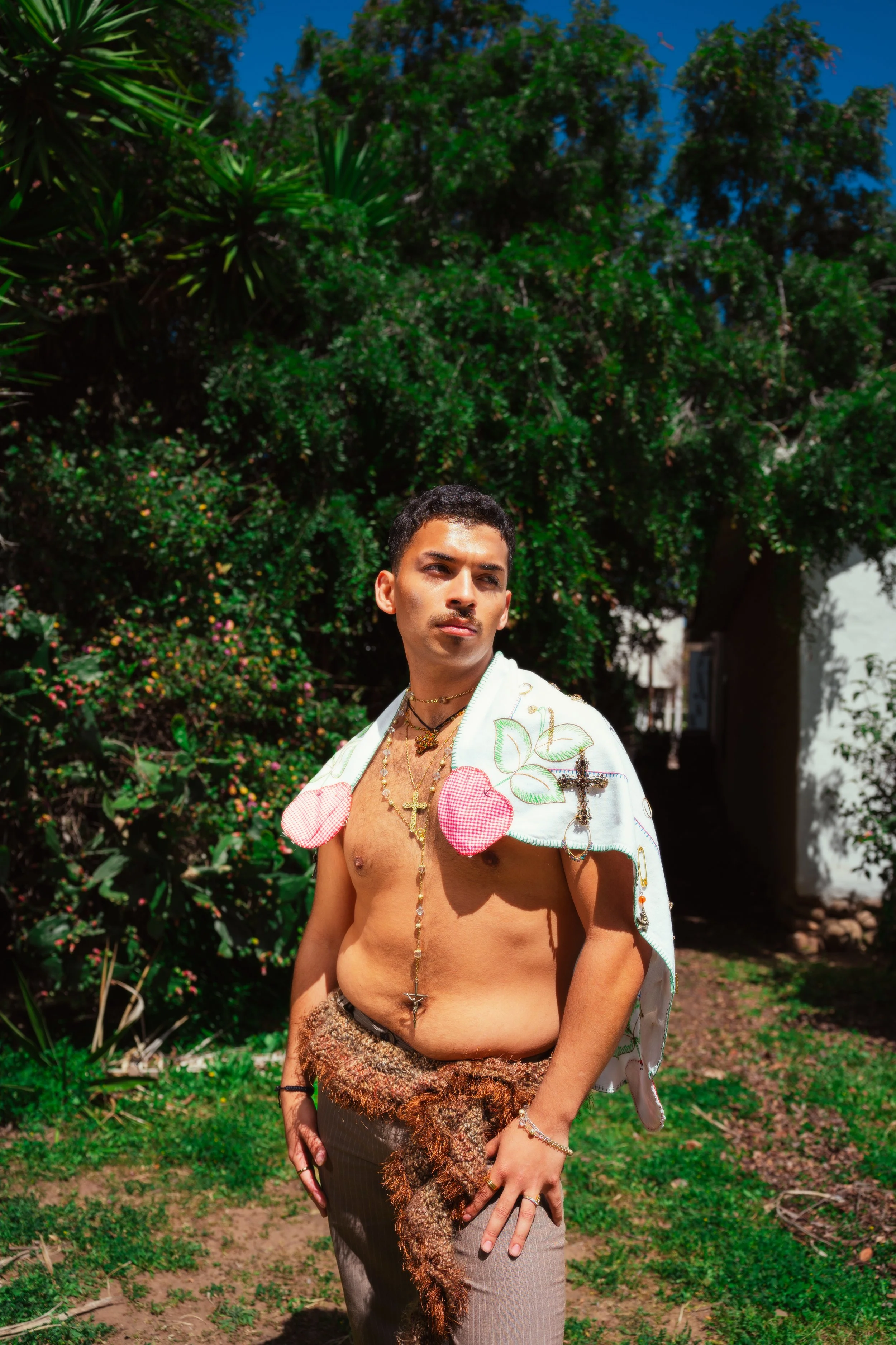 A young man standing outdoors in front of greenery, shirtless with a white embroidered cloth draped over his shoulders, wearing jewelry and a fur wrap around his waist.
