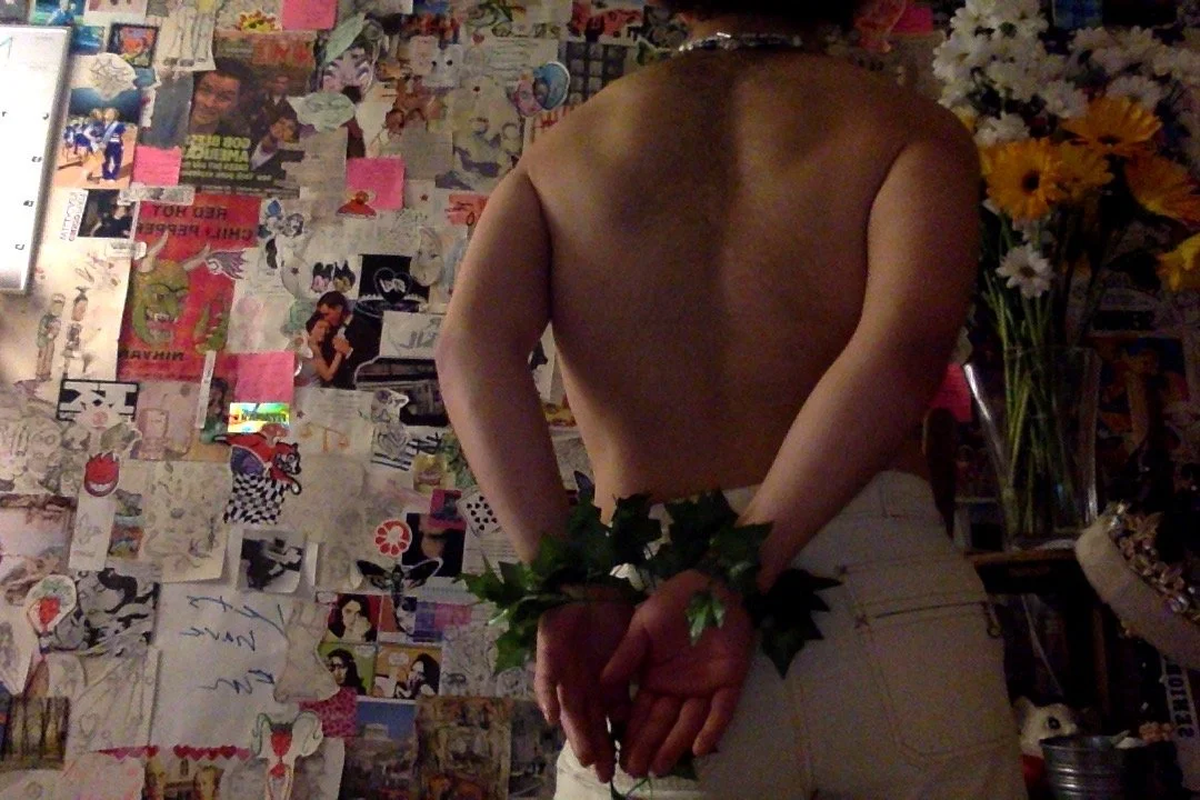A person with no shirt, holding leafy greenery behind their back with both hands, standing in front of a wall covered with posters and pictures, with a bouquet of flowers visible on the right side.