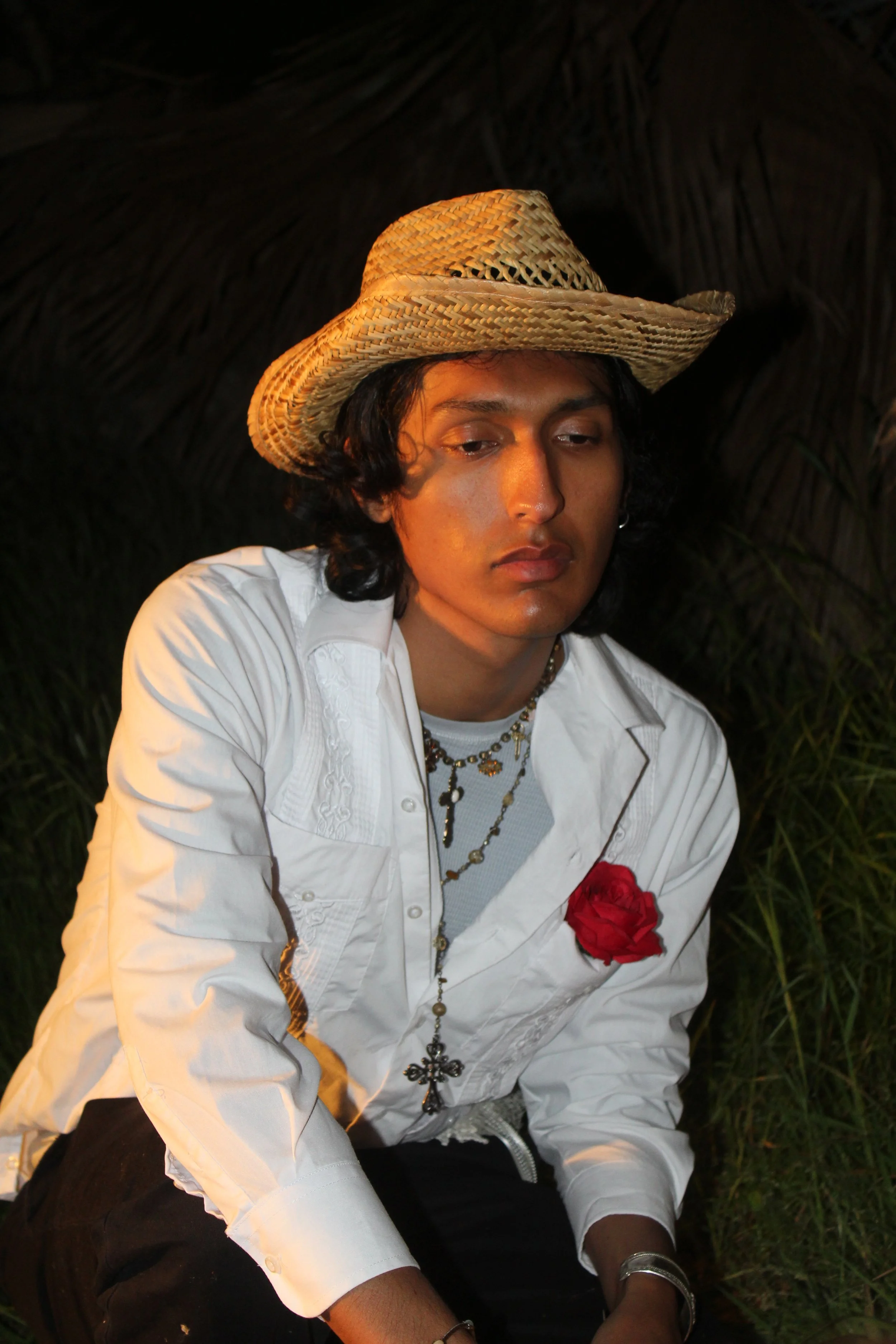 A young man with long dark hair, wearing a straw hat, a white embroidered shirt with a red rose pinned to it, layered necklaces, and a bracelet, sitting outdoors with tall grass and a dark background.