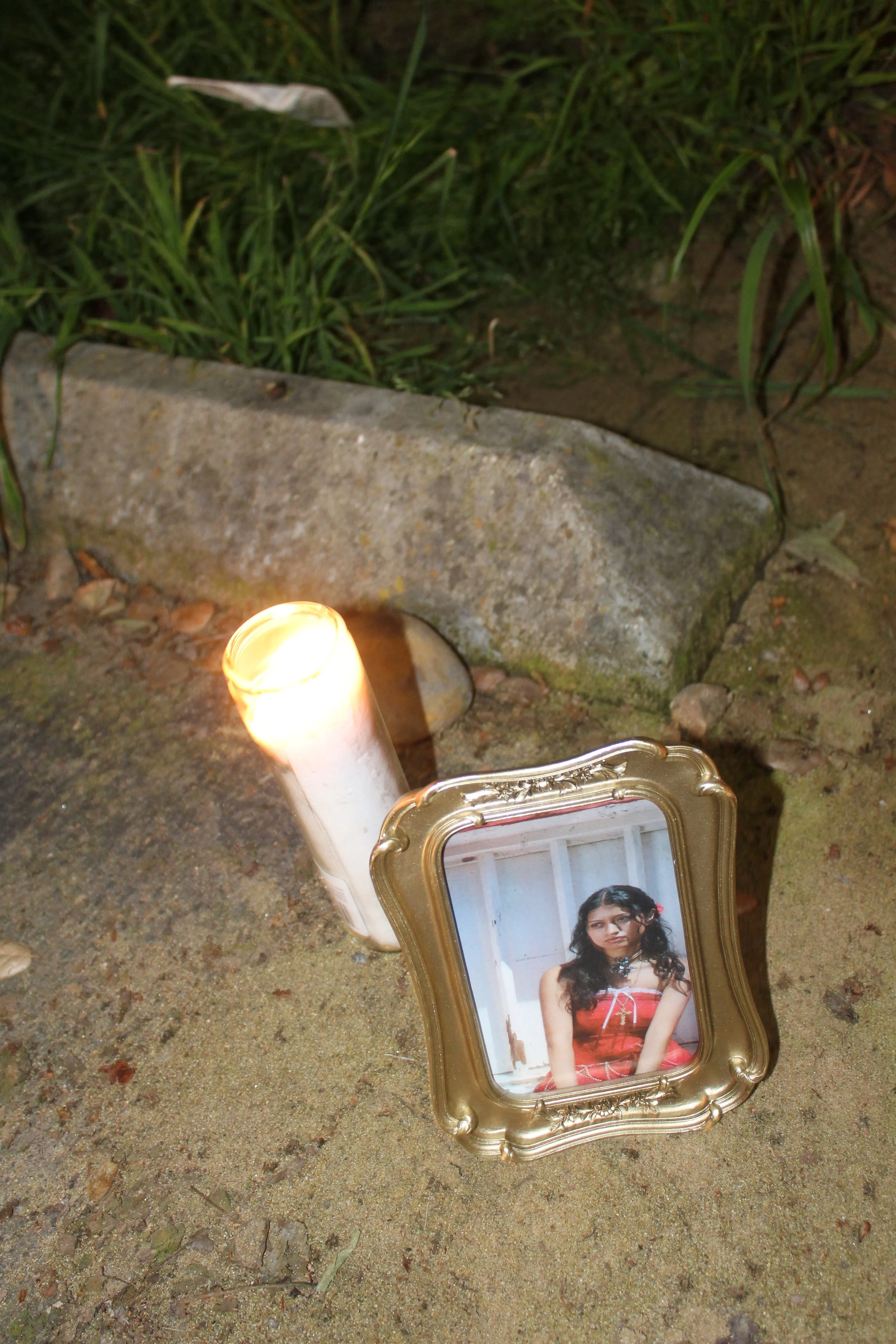 A lit candle and a small ornate framed photograph of a woman in a red dress placed on sandy ground outdoors, with grass and rocks in the background.