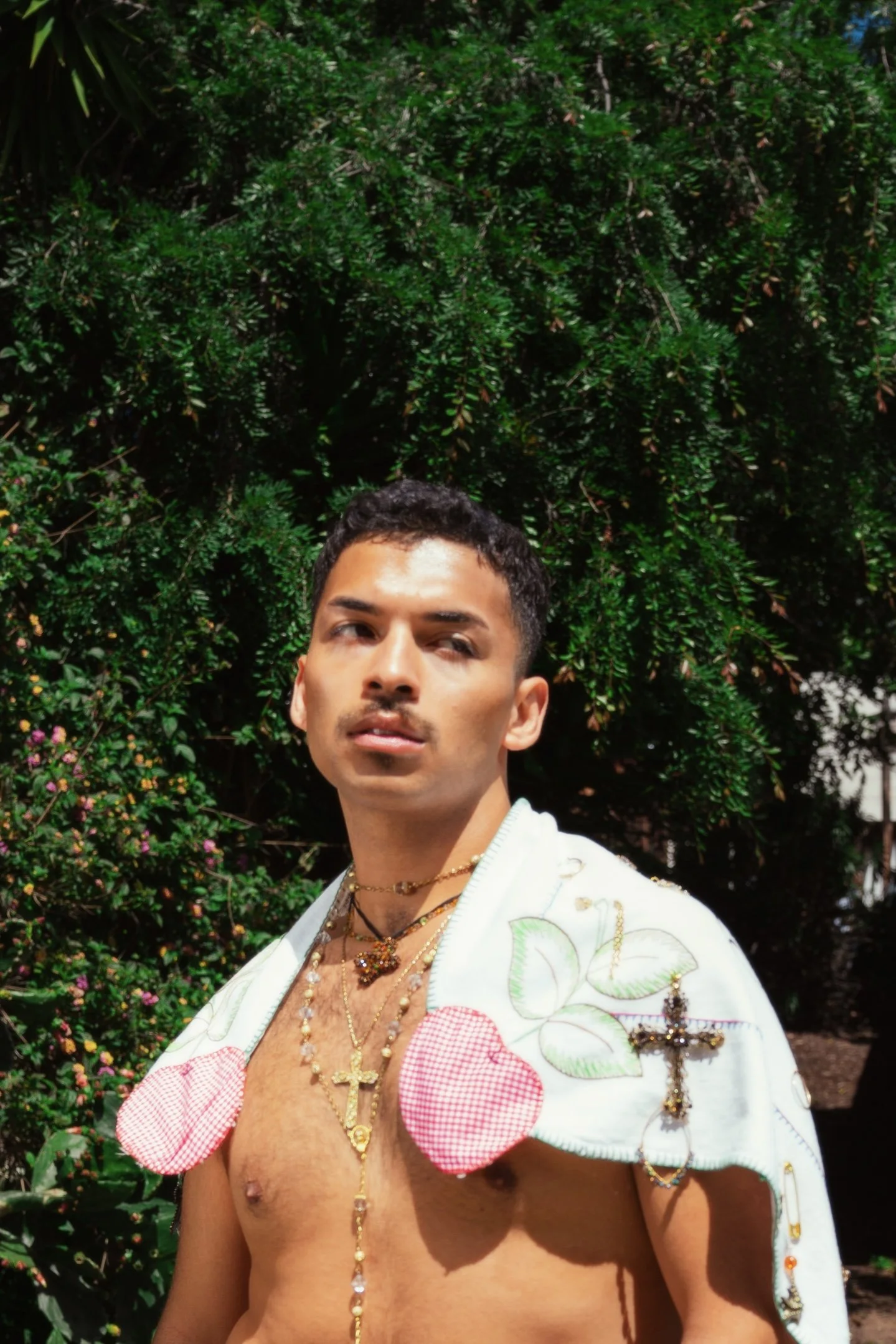 A young man standing outdoors with a large green bush behind him, wearing multiple gold necklaces and a white embroidered shawl with red checkered hearts and green leaves.
