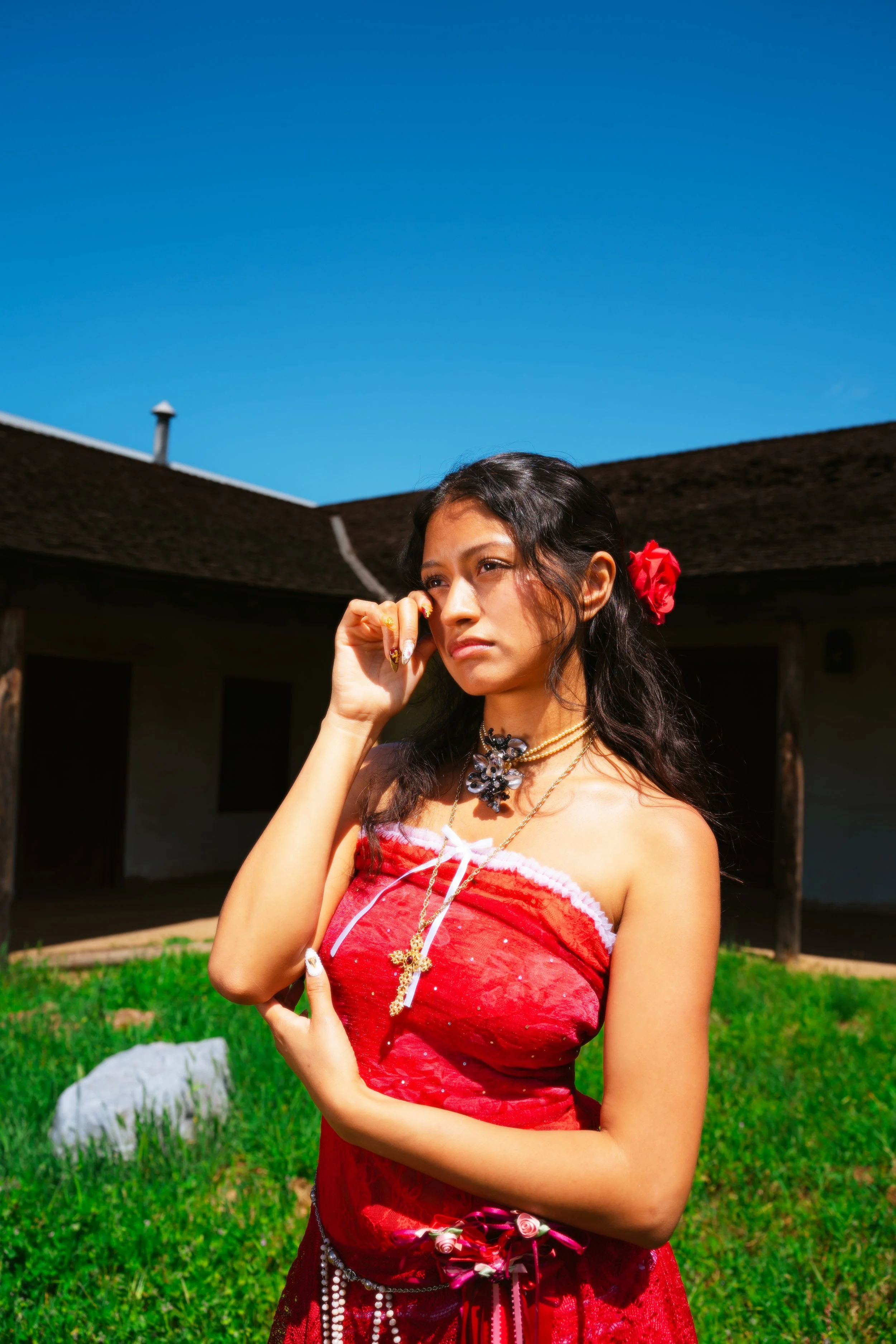 A woman in a red strapless dress with jewelry and a flower in her hair stands outside under a blue sky.
