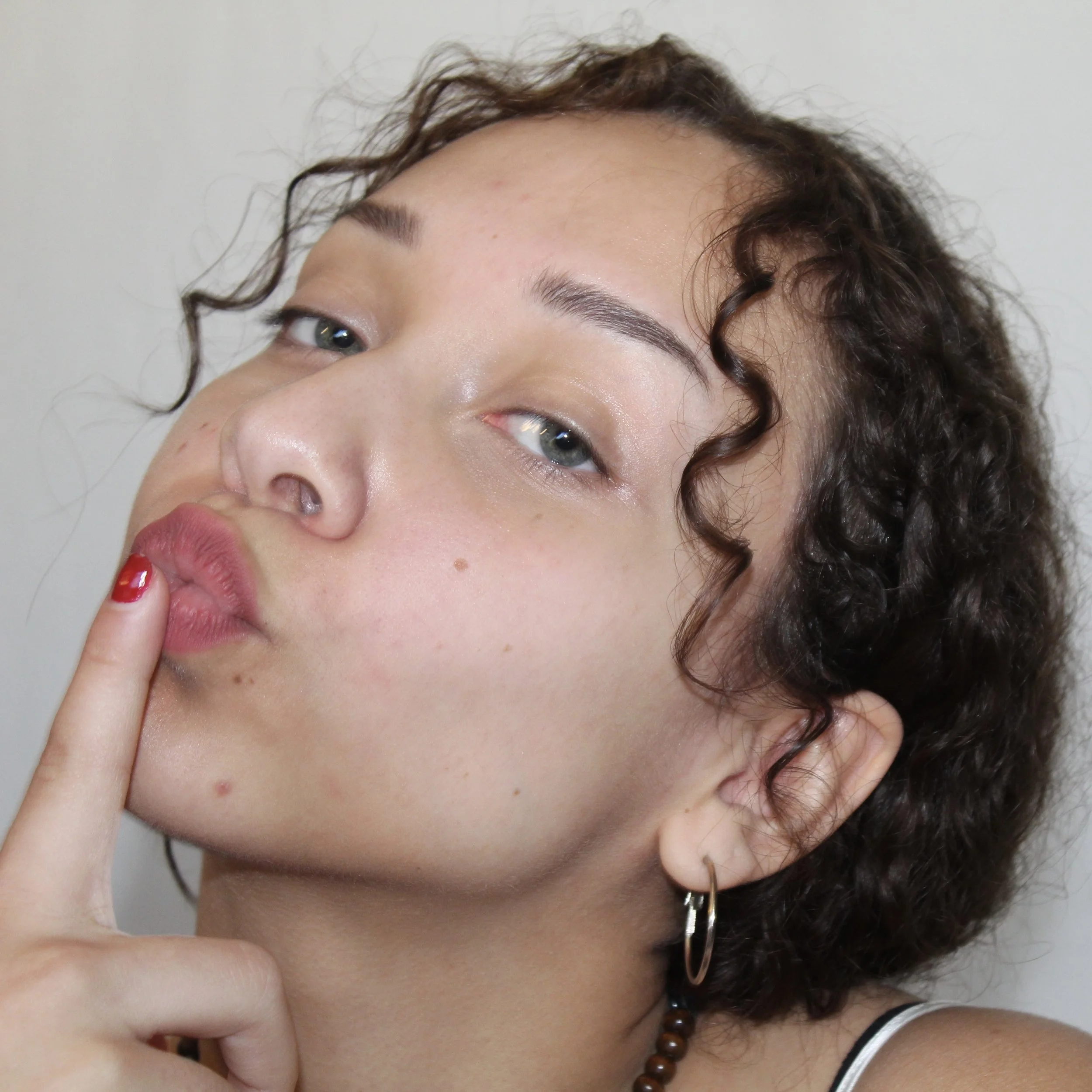 A close-up of a woman with curly hair making a pouting face and holding her finger to her lips.