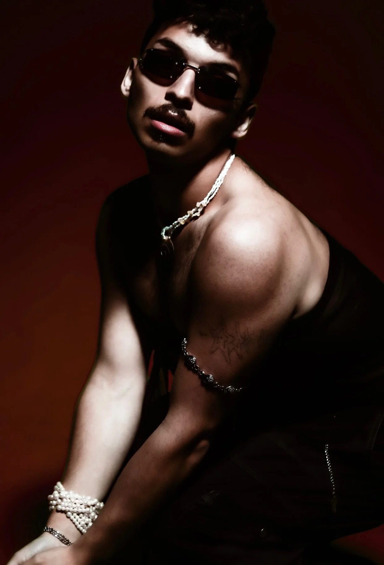 A young man with dark hair, wearing black sunglasses, a necklace, and jewelry on his wrist and arm, posing shirtless against a dark red background.