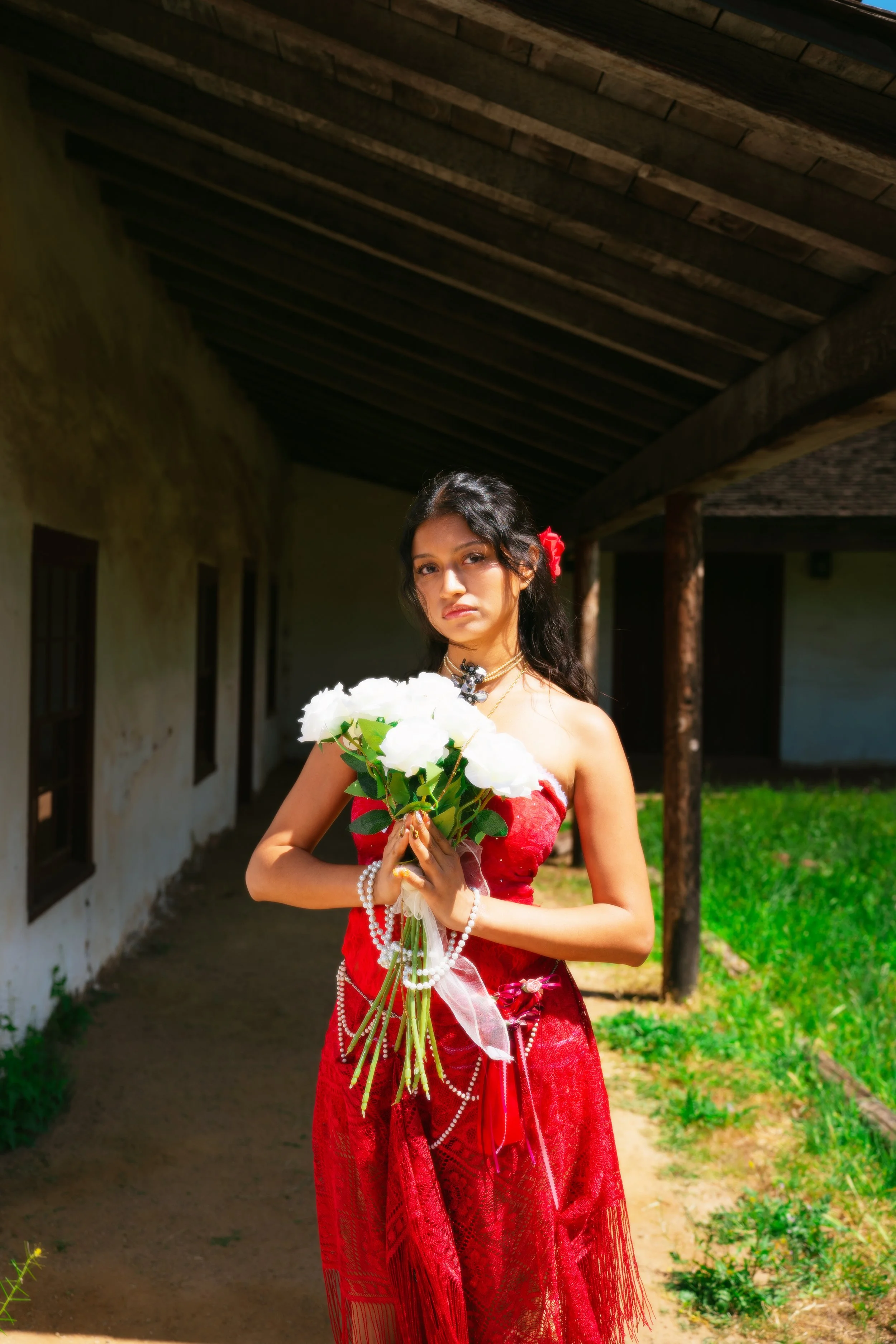 A woman in a red traditional dress holding a bouquet of white flowers, standing outdoors near an old building with wooden beams and windows.