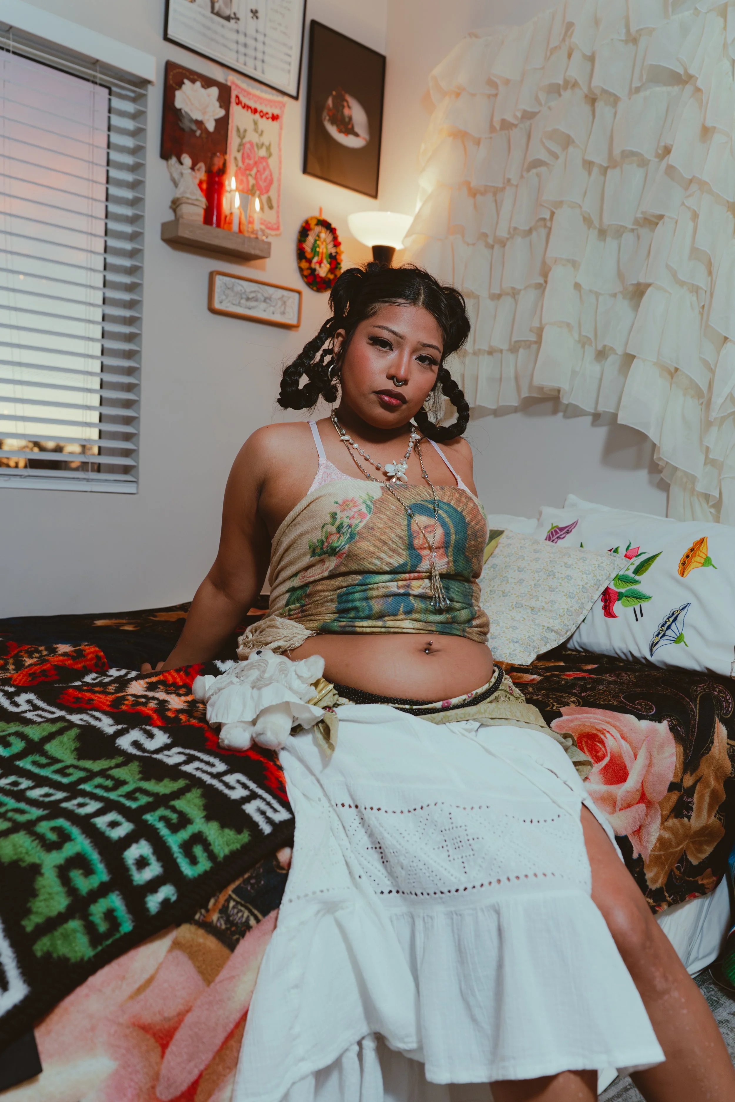 A young woman with dark hair styled in braids sitting on a bed in a room with various wall decorations. She wears a colorful crop top, jewelry, and a white skirt, with stuffed animals and pillows around her.