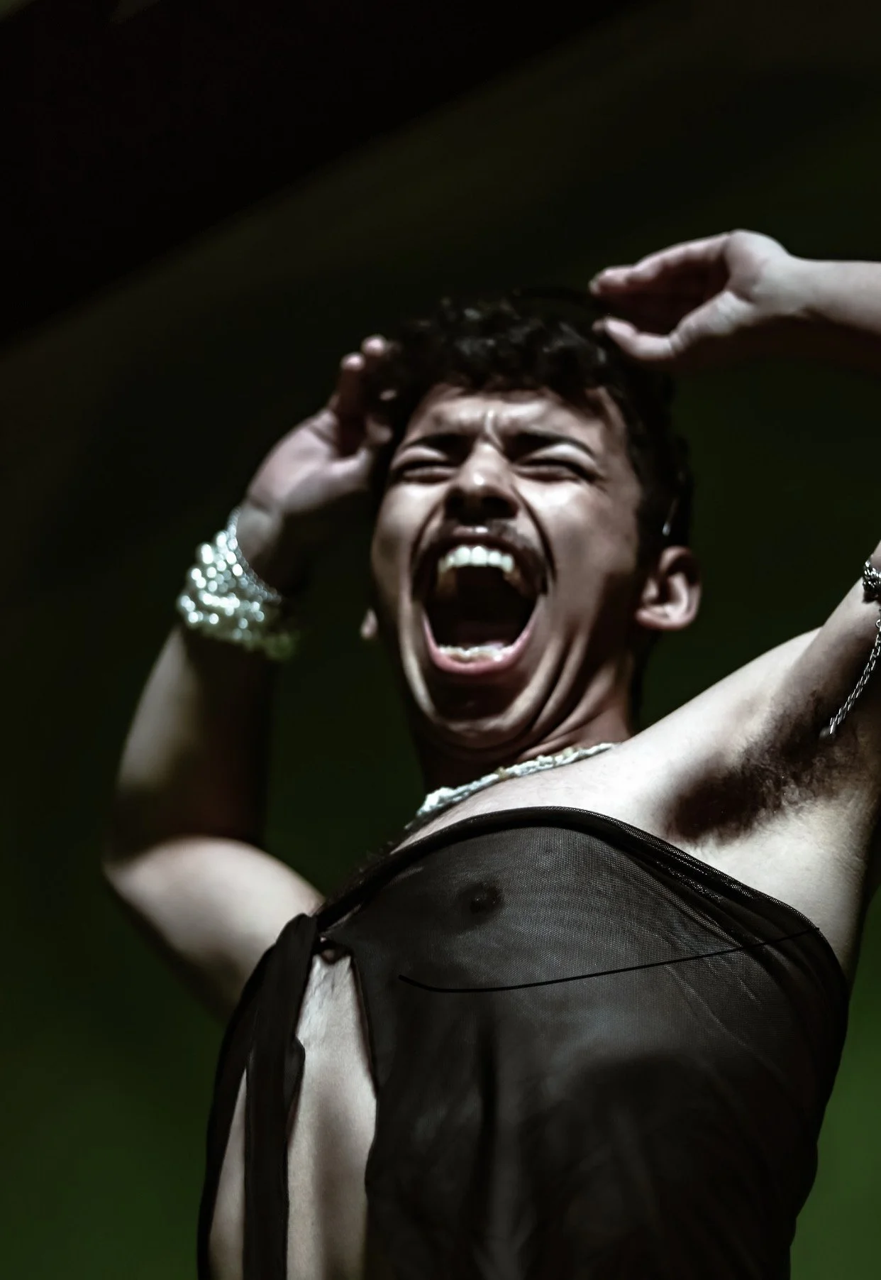 A person with short curly hair and dark skin, wearing a black mesh top, is captured in a moment of intense emotion, with their mouth wide open in a scream or shout, arms raised, and eyes closed.