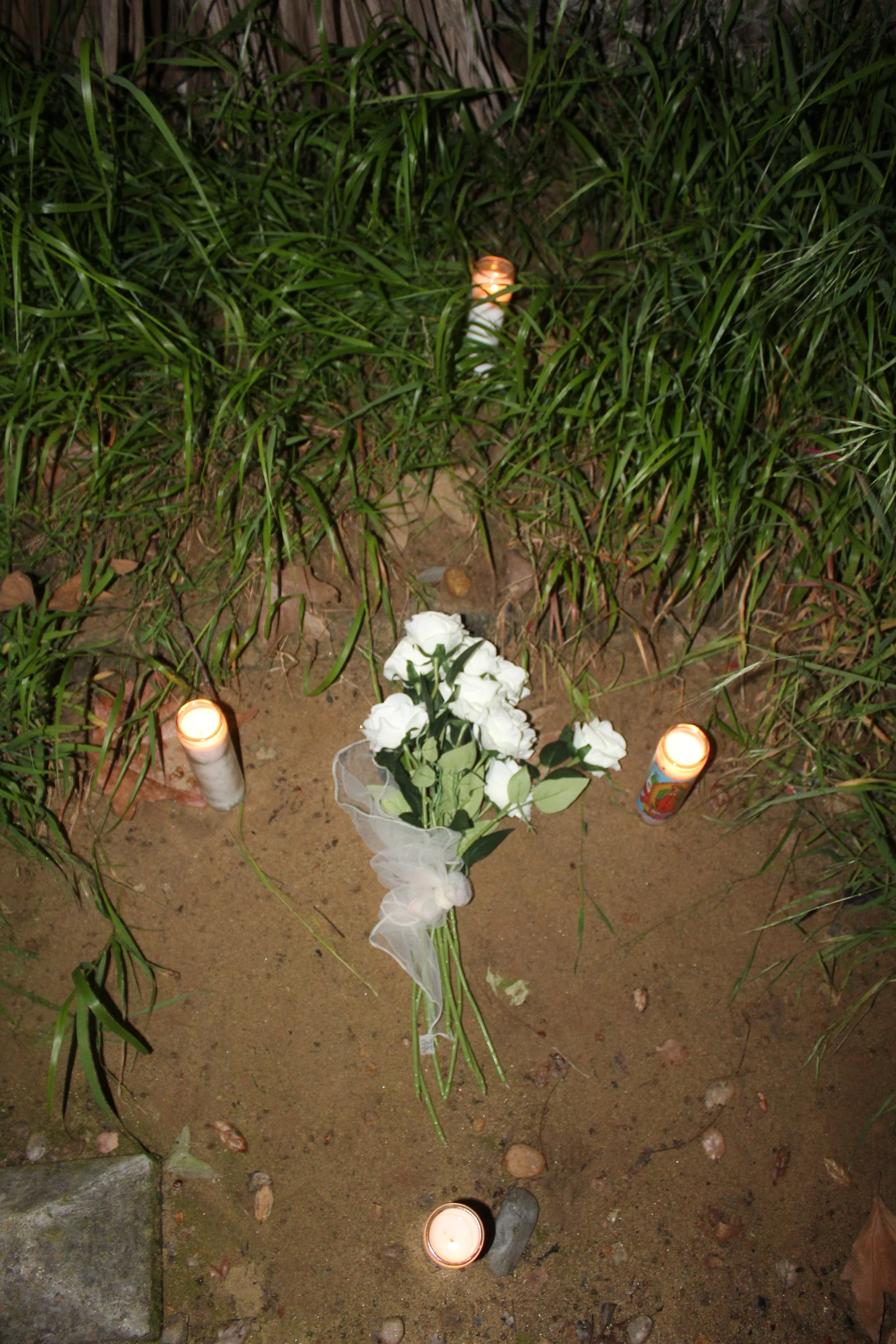A memorial site with white flowers and four lit candles on the ground surrounded by grass and dirt.