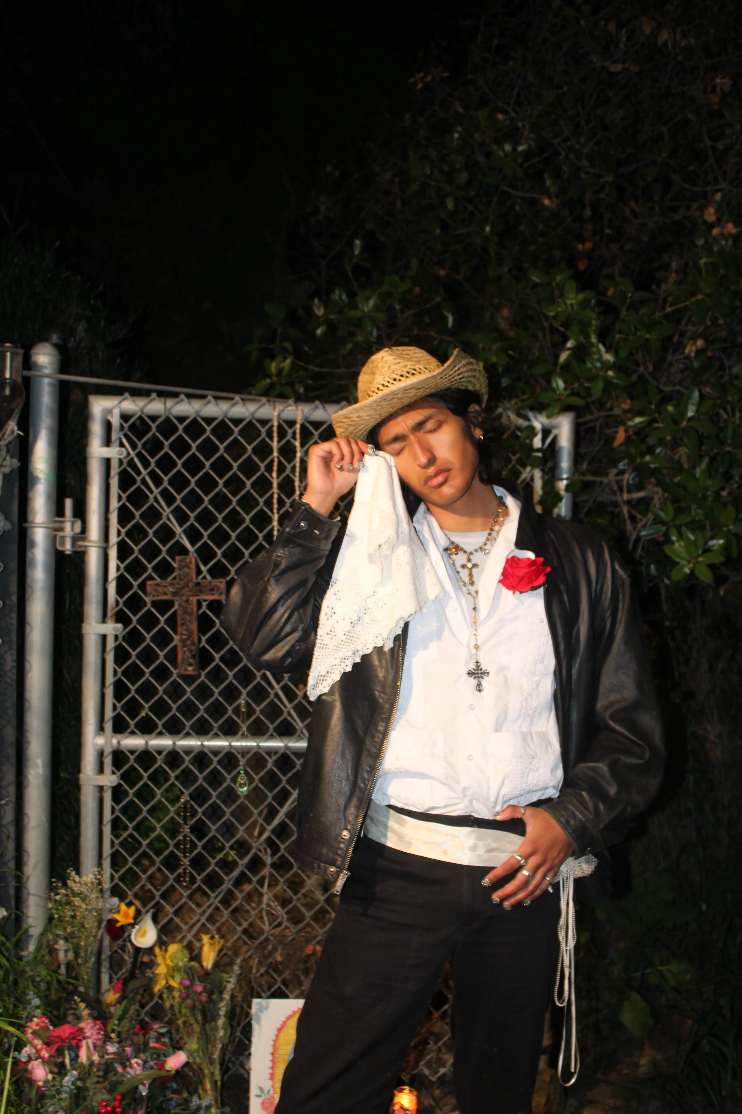 A young person wearing a straw hat, white shirt with lace, black leather jacket, and black pants, standing in front of a memorial with flowers and a small cross, holding a tissue to their face in a sorrowful moment.