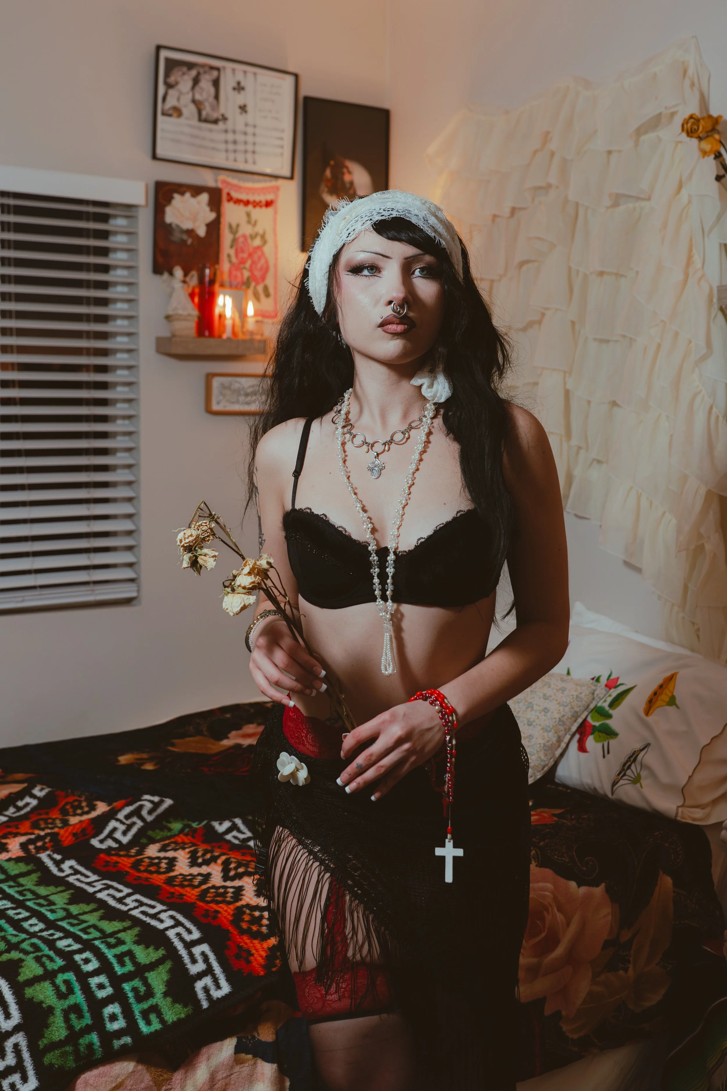 A woman with black wavy hair and makeup, wearing a white lace head covering, black lace bra, and black fringed skirt, holding dried flowers and a rosary with a cross, standing in a decorated bedroom with religious and floral artwork on the wall.