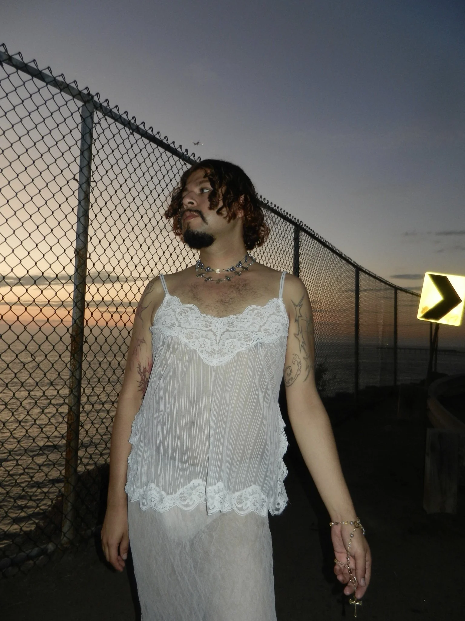 A person with curly hair, tattoos, and jewelry wearing a white lace and sheer dress, standing near a chain-link fence at sunset, with an airplane and directional sign in the background.