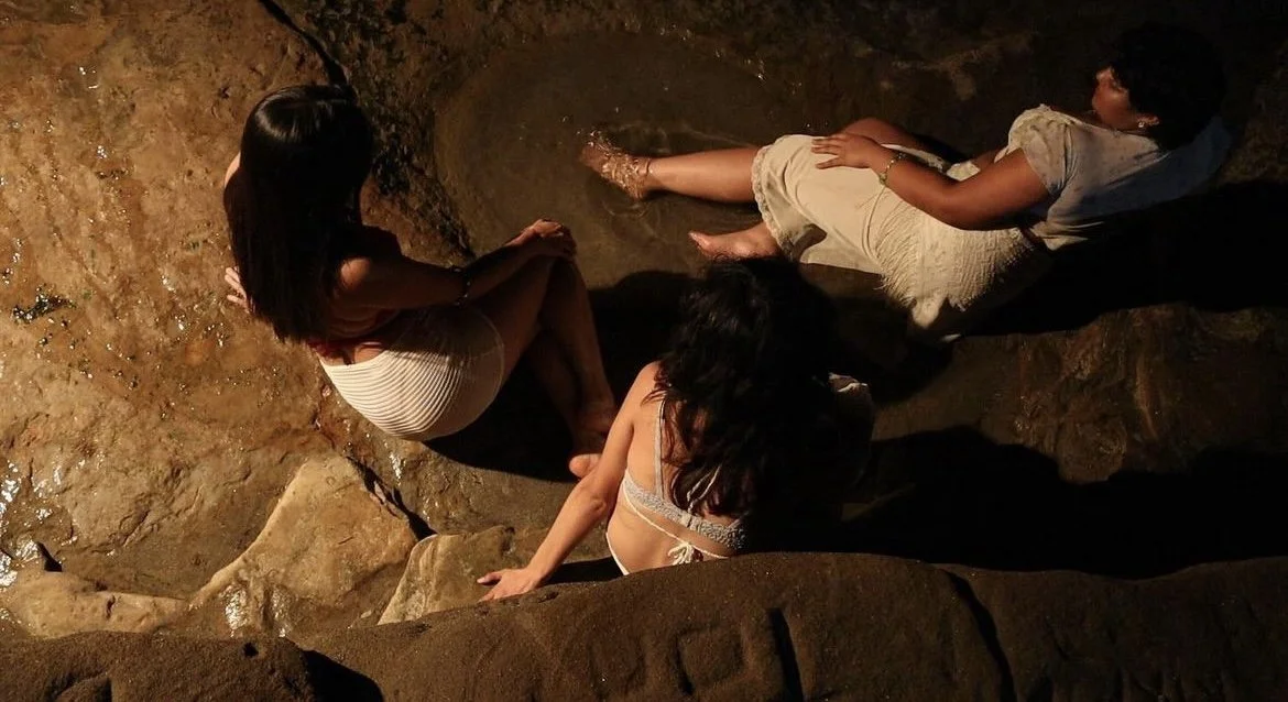 Three women sitting on rocks near a shallow water body in a dark, natural setting.