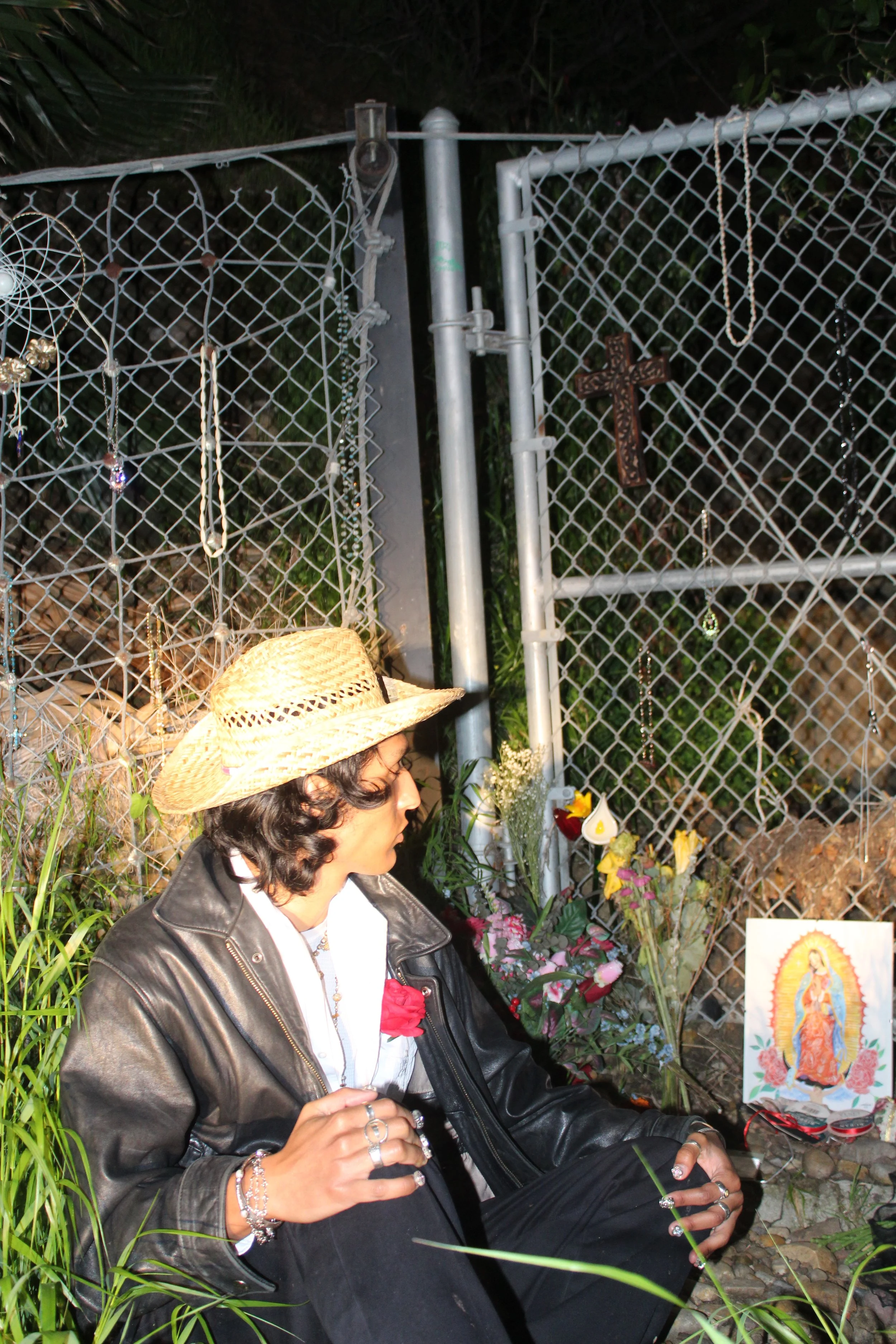 A person wearing a straw hat and black leather jacket sitting by a makeshift memorial with flowers, a religious icon, and a wooden cross, next to a chain-link fence at night.