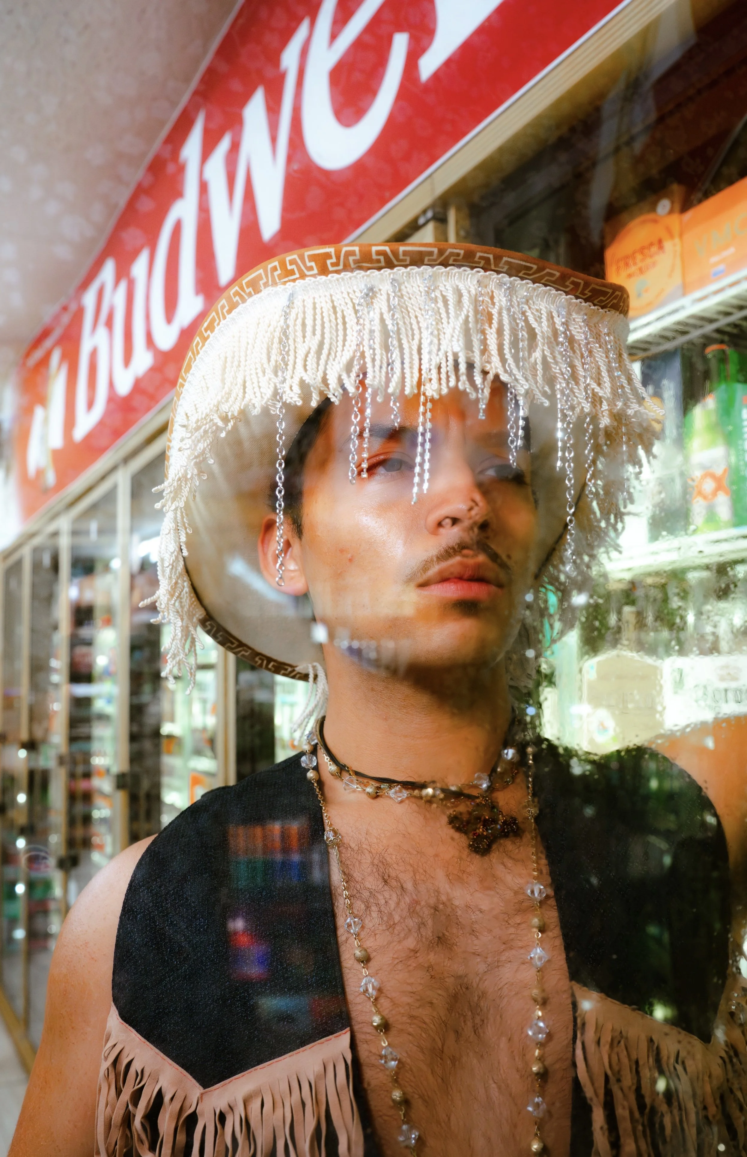 A young man with dark hair and sparse facial hair wearing a large, decorative hat with beads hanging from it, a black vest with fringes, and layered necklaces, standing behind a glass window in a convenience store with a red and white sign in the bac