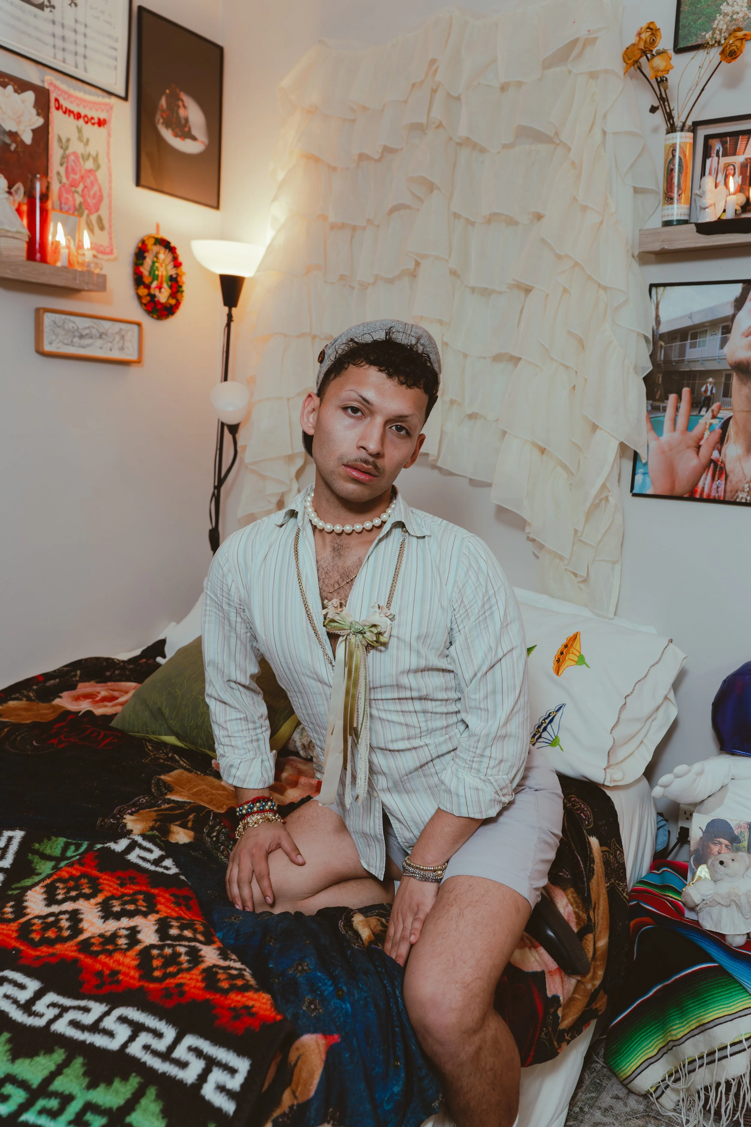 A young man with curly dark hair, wearing a baseball cap, a pearl necklace, and a striped shirt, kneels on a bed in a decorated room. The room features various wall decorations, a light lamp, and personal items on shelves. The bed has a colorful blan