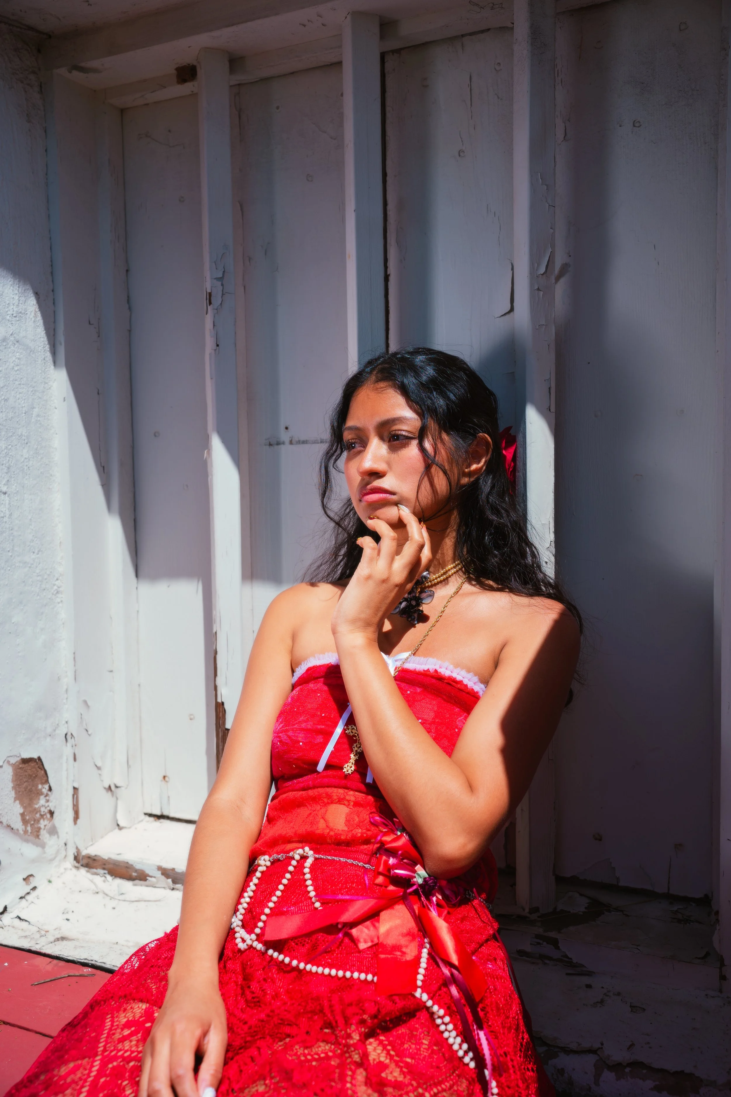A young woman with dark wavy hair and a red flower in her hair, wearing a red dress with lace and pearl accents, sits against a weathered white wooden wall, looking contemplative.