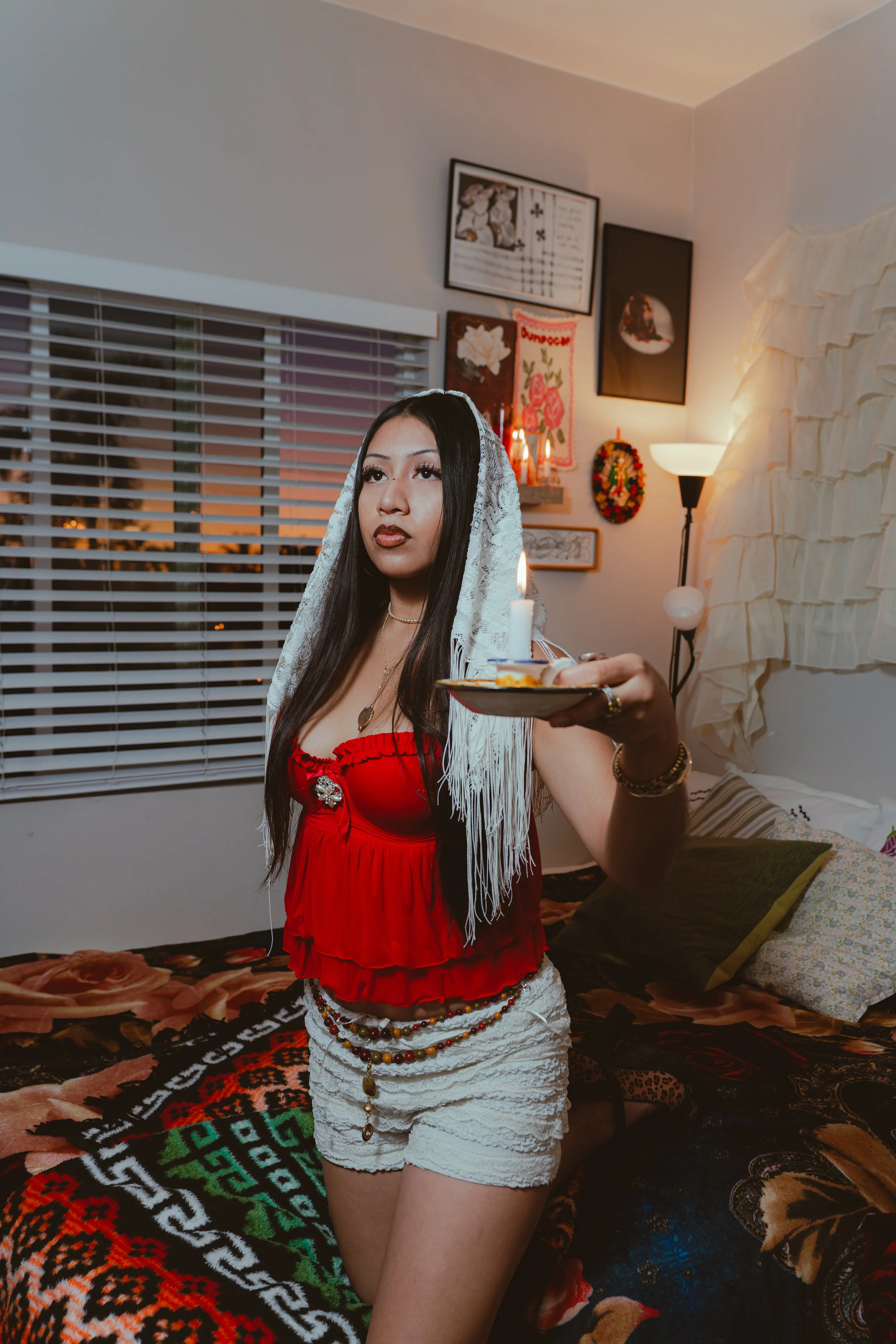 A woman wearing a red top and white shorts holding a tray with a lit candle in a bedroom with various artwork on the wall and a window with blinds.