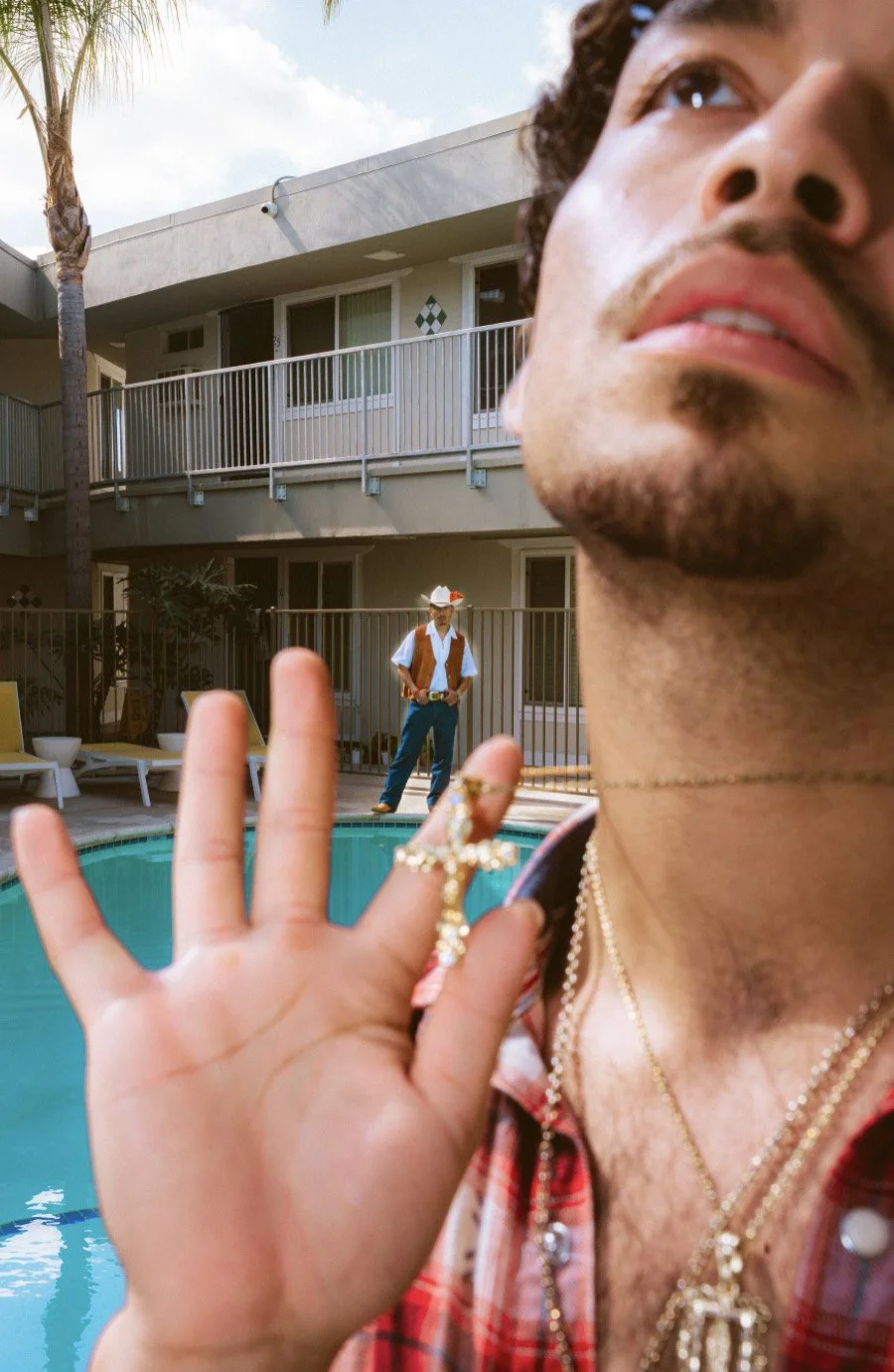 Close-up of a man's face and hand with jewelry, with a swimming pool and a person dressed as a cowboy in the background.