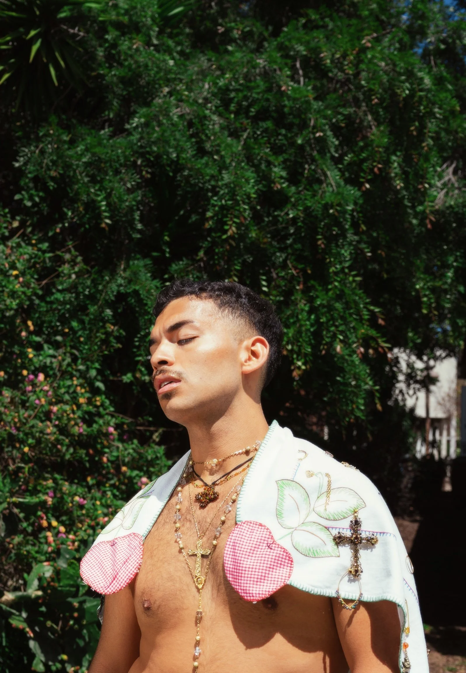A shirtless man wearing multiple layered necklaces, including a cross and a floral pendant, with a white embroidered cape featuring pink checkered patches and green leaf designs, standing outdoors in front of lush green foliage.