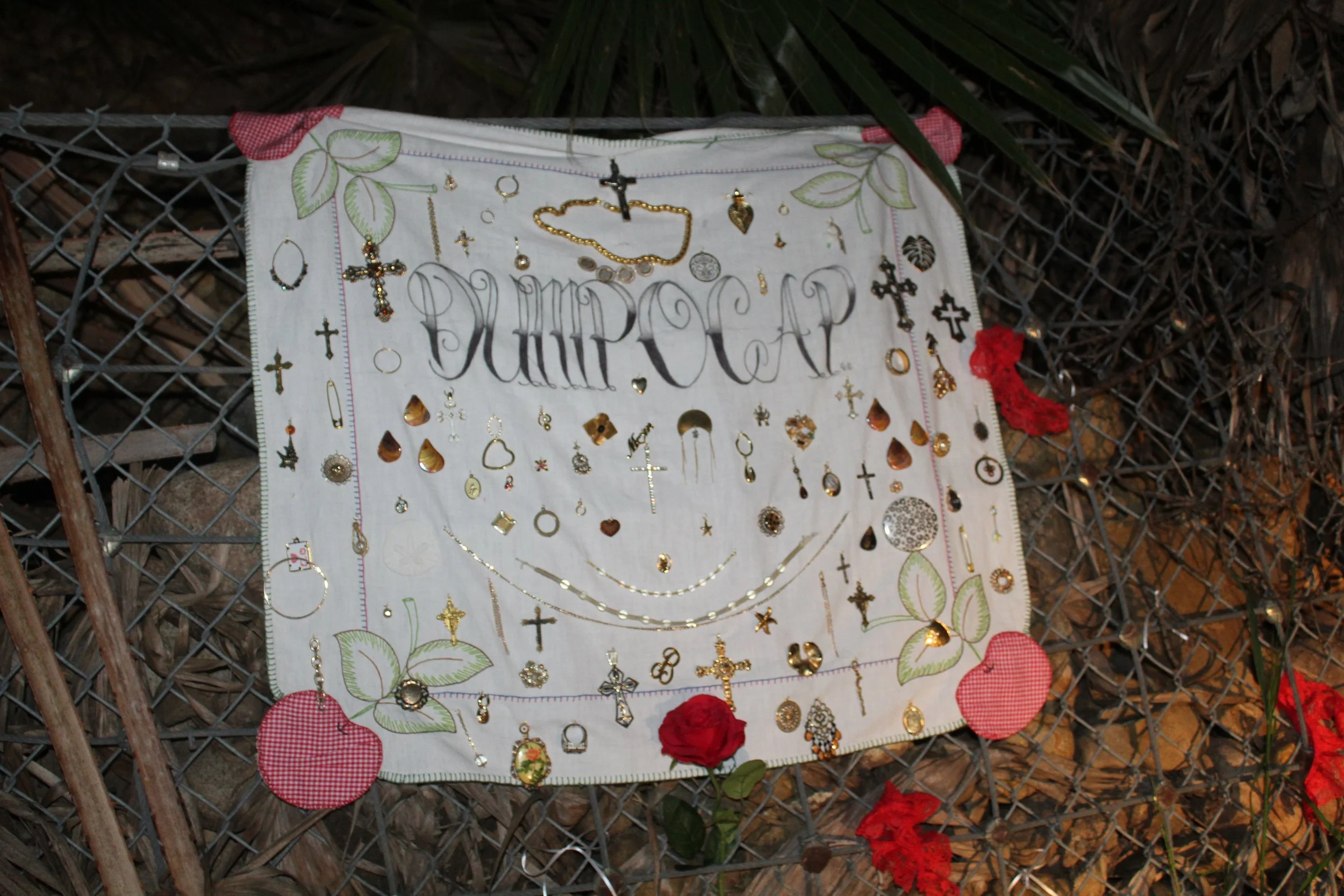 A fabric memorial with the words 'DUMP CAMP' embroidered on it, decorated with various jewelry pieces, crosses, and symbols, and bordered with red and checkered fabric, attached to a wire fence with red flowers nearby.