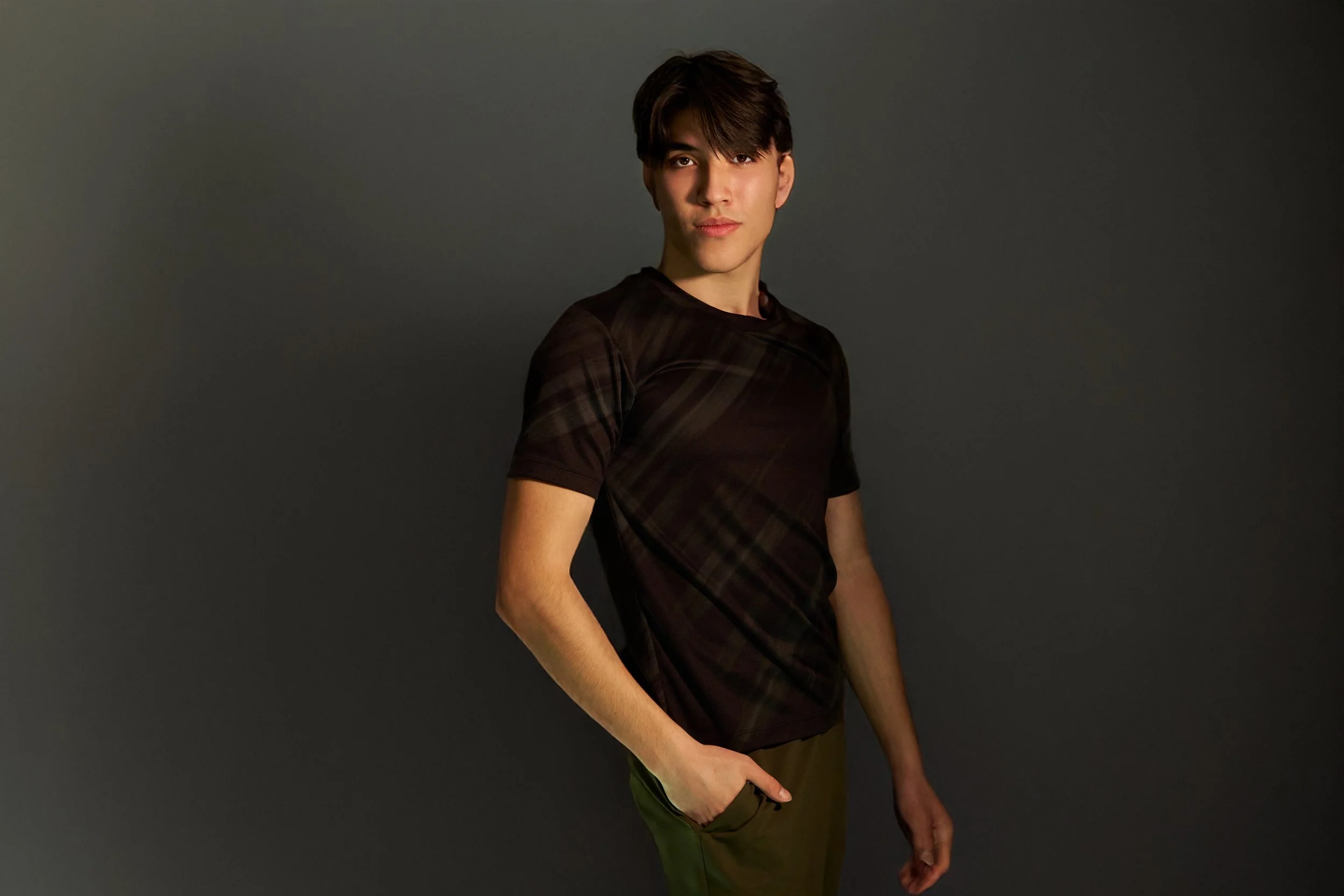 Young man with dark hair wearing a black t-shirt and green pants standing against a dark gray background.
